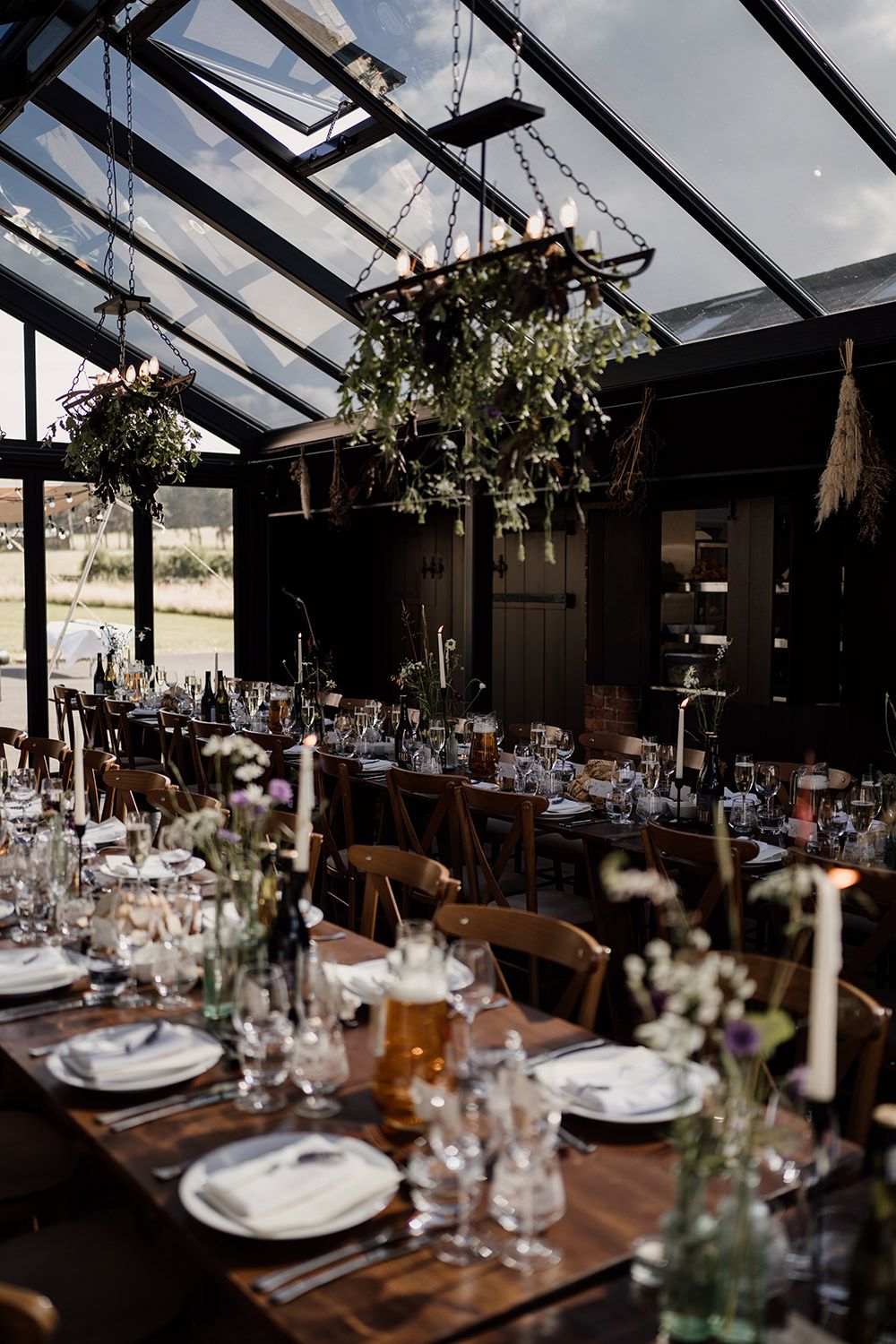 Long tables styled and ready for wedding guests with drinks, flowers, lit with natural light in a glass building.
