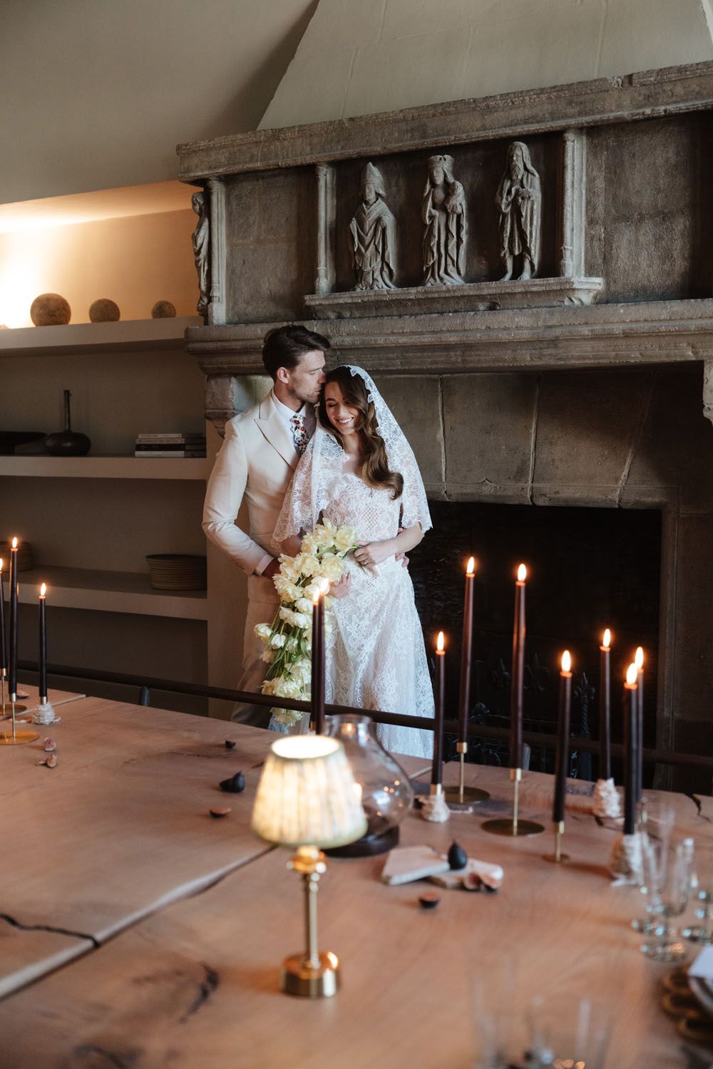 Bride and groom embrace by a stone fireplace with candles and floral bouquet on a rustic wooden table.