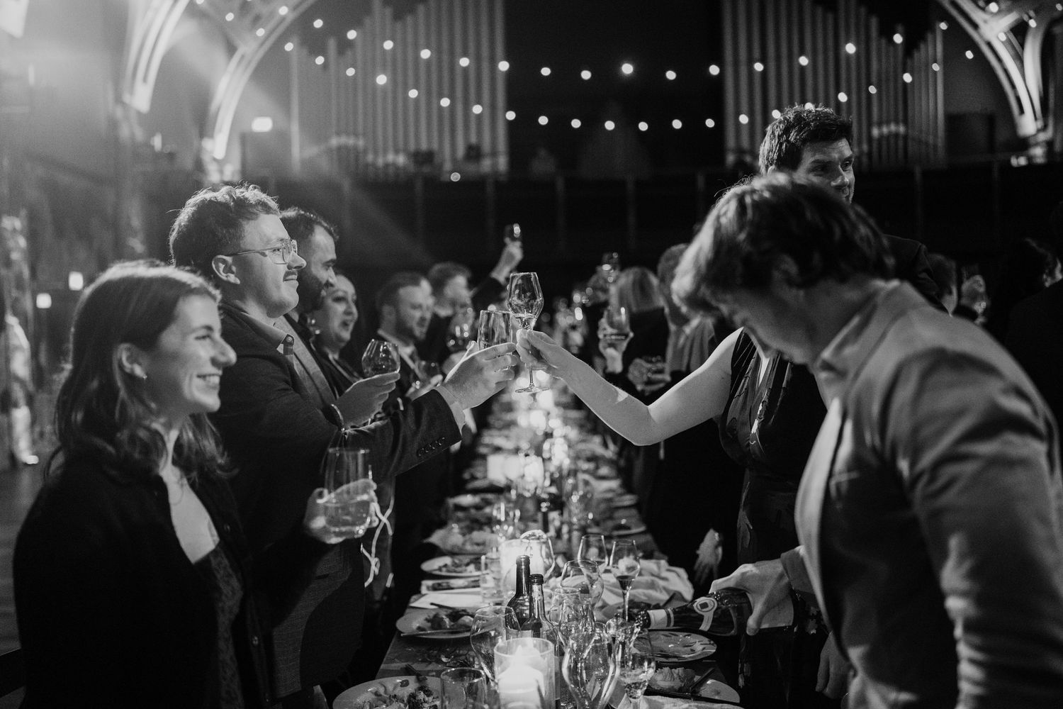 Black and white photo of wedding guests toasting at candlelit long reception dinner table.