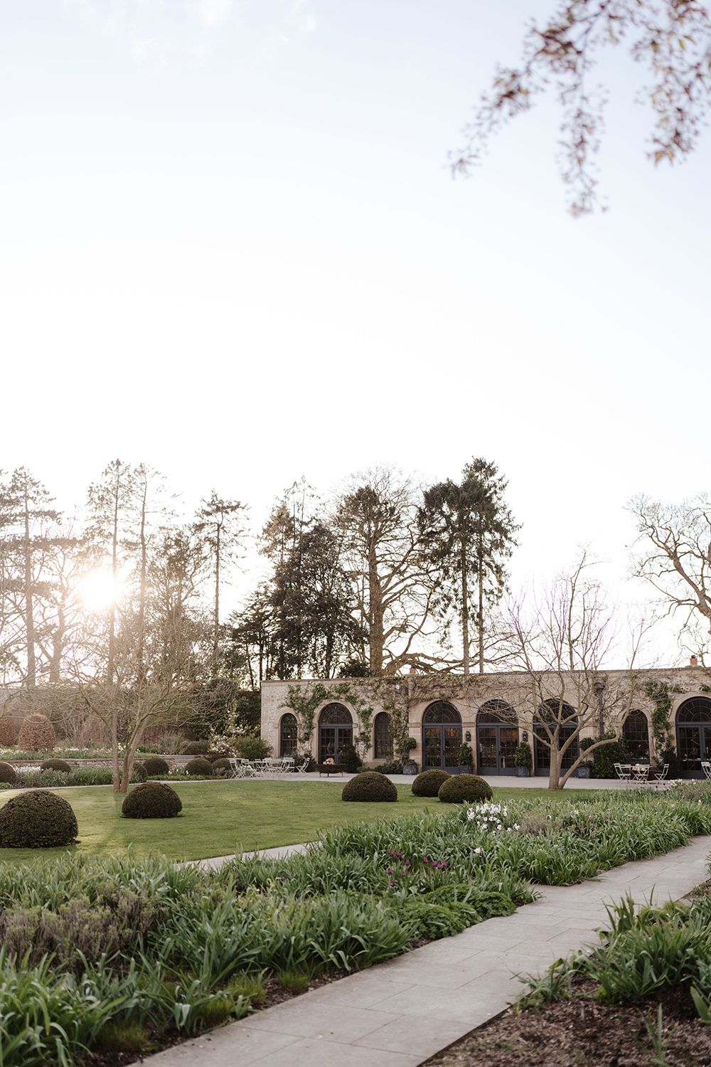 Stone arched walkway in a formal garden with manicured hedges and paths at sunset through towering pine trees.