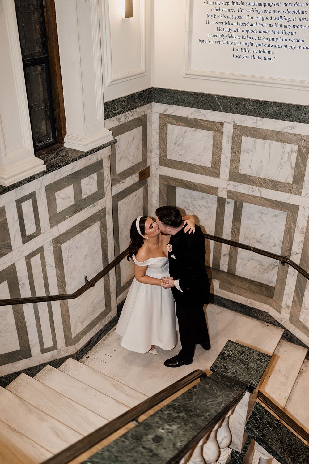 Couple in wedding attire share an intimate moment on elegant marble stairs with geometric wall patterns.