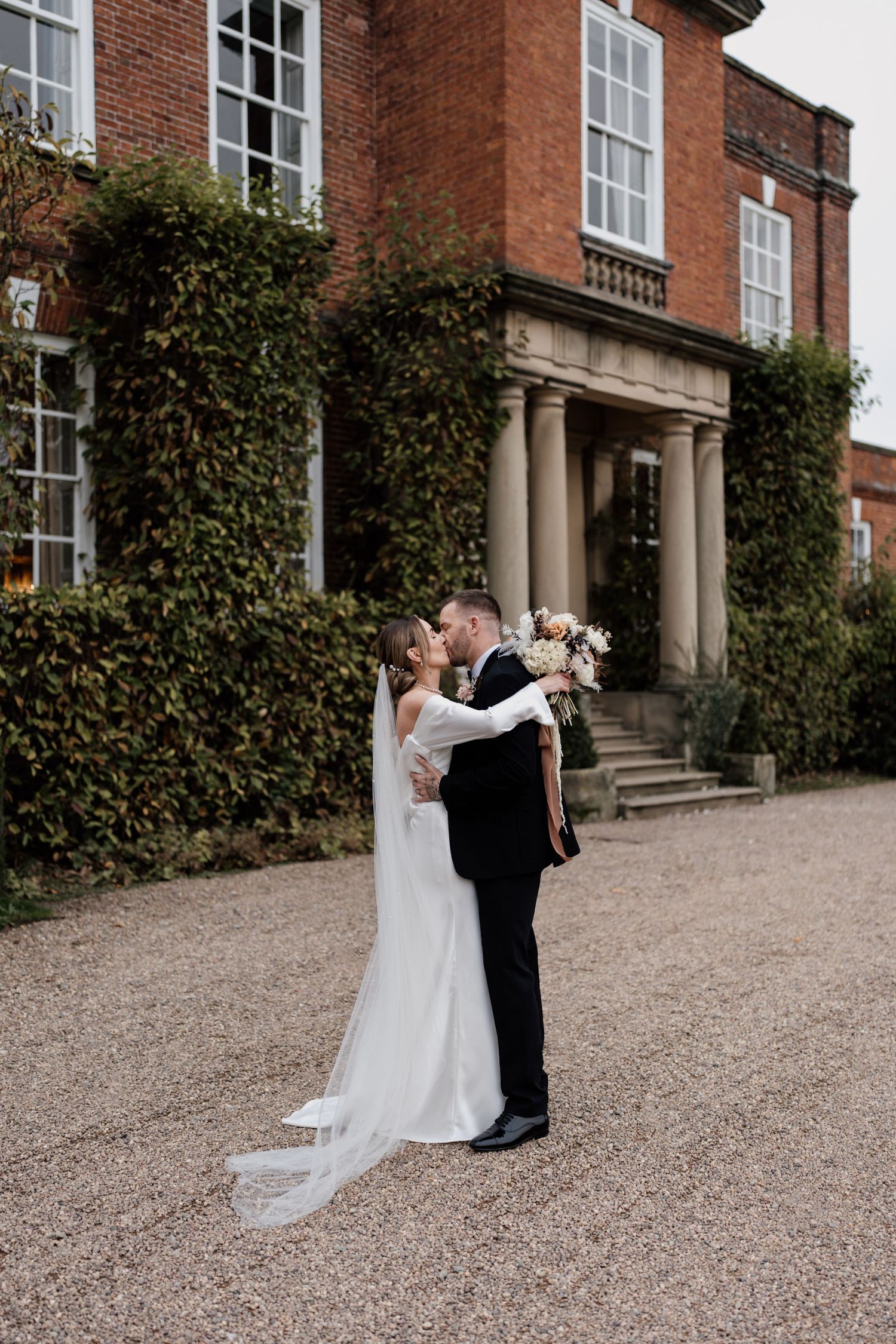 Couple embraces outside a grand brick Georgian manor house with climbing ivy and white columns on their wedding day.
