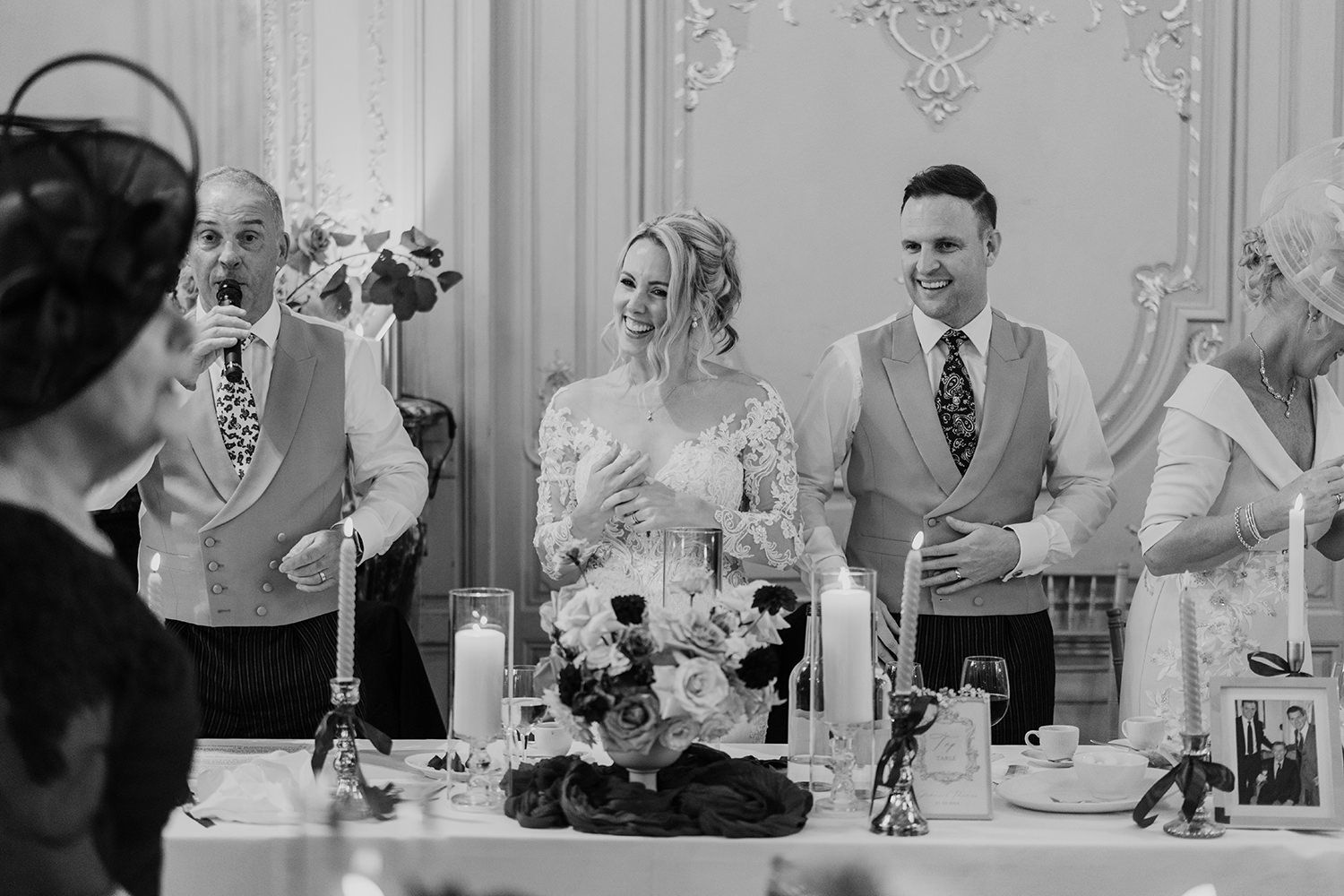 Wedding guests raise their glasses in celebration at an elegant monochrome reception with ornate decor and floral centerpieces.