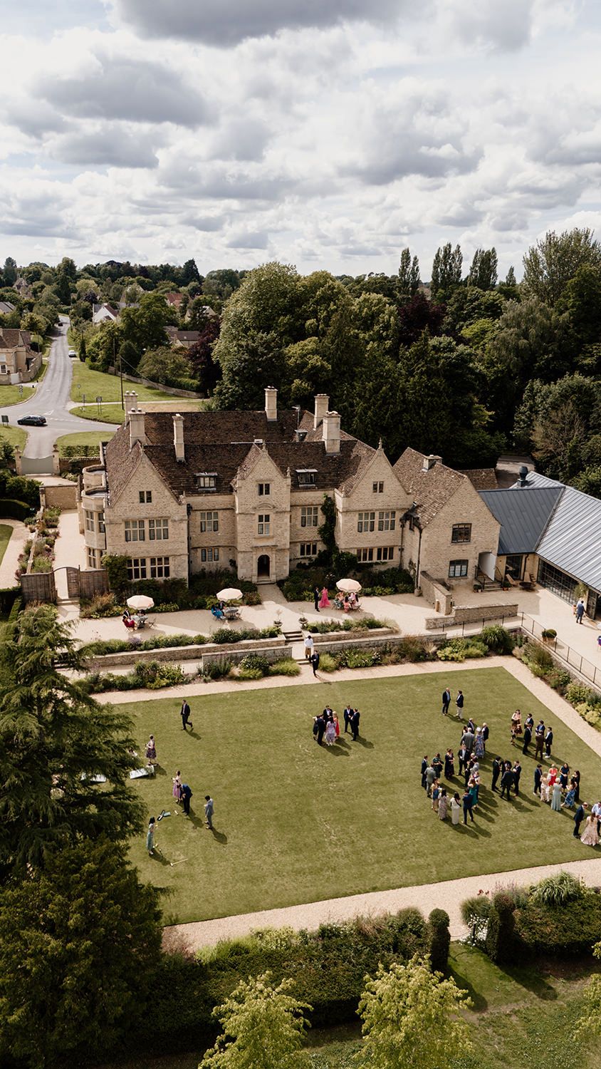 Aerial view of a grand English manor house with manicured gardens filled with wedding guests on a sunny day.