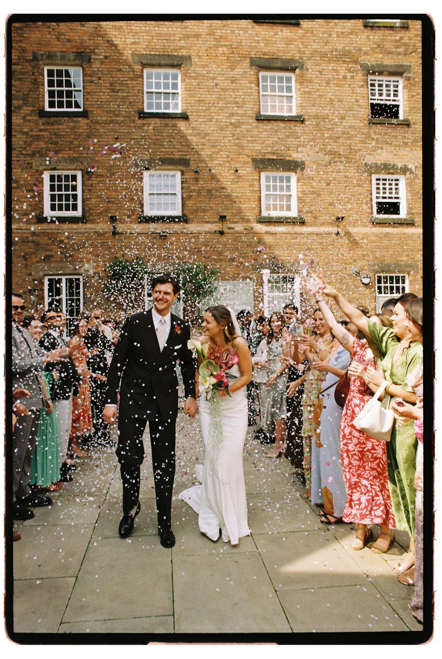 Bride and groom walk through confetti shower with smiling wedding guests outside a brick building.