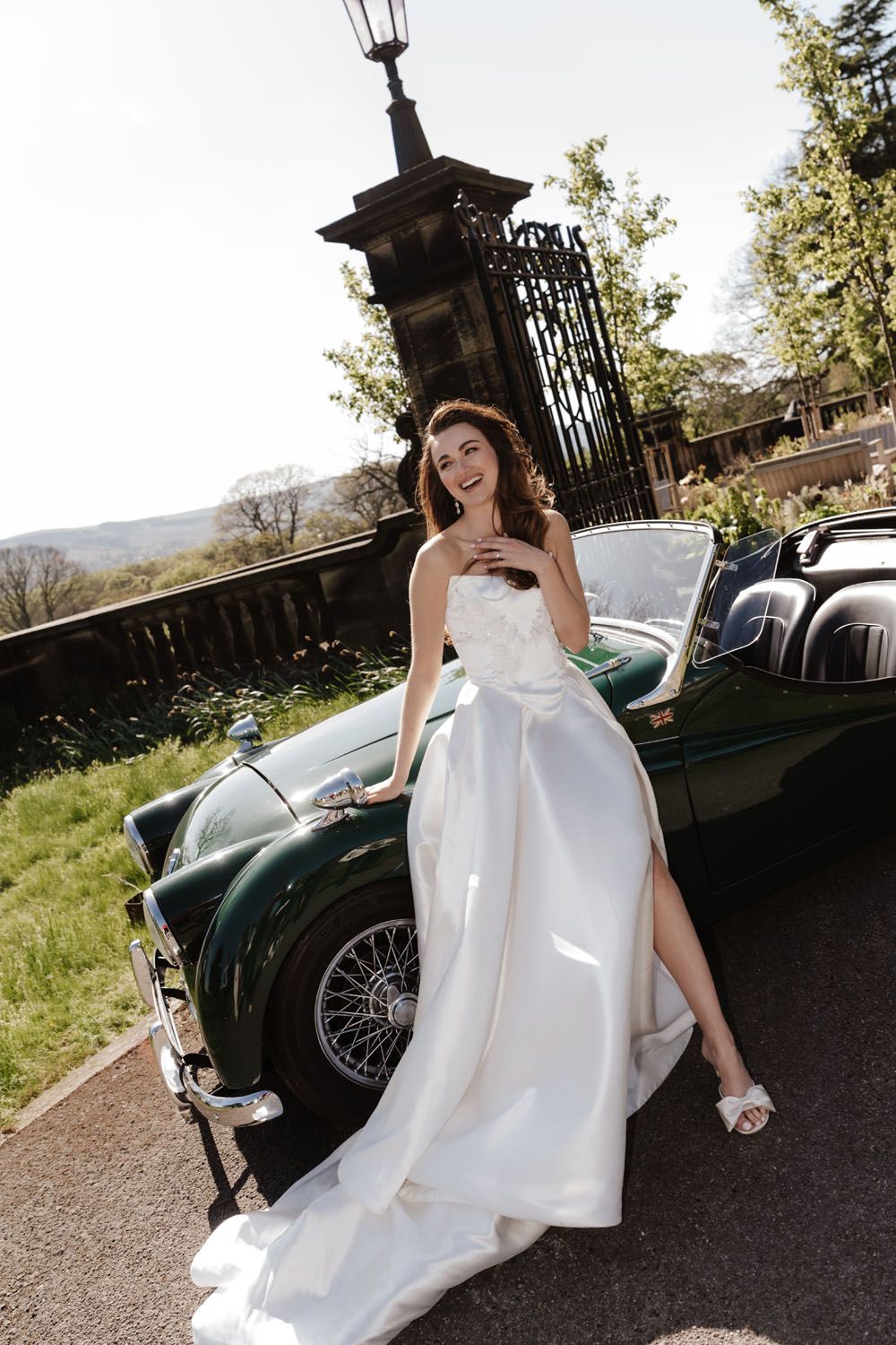 Smiling bride in white gown leaning against a vintage green convertible car outdoors on a sunny day.