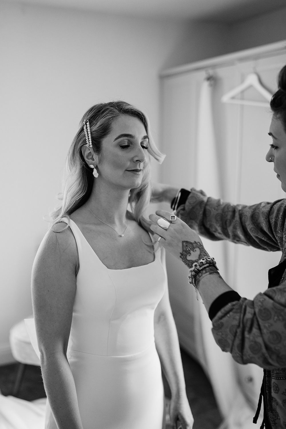 Bride stood with eyes closed in her wedding dress while friend adjusts her straps. Room is behind.