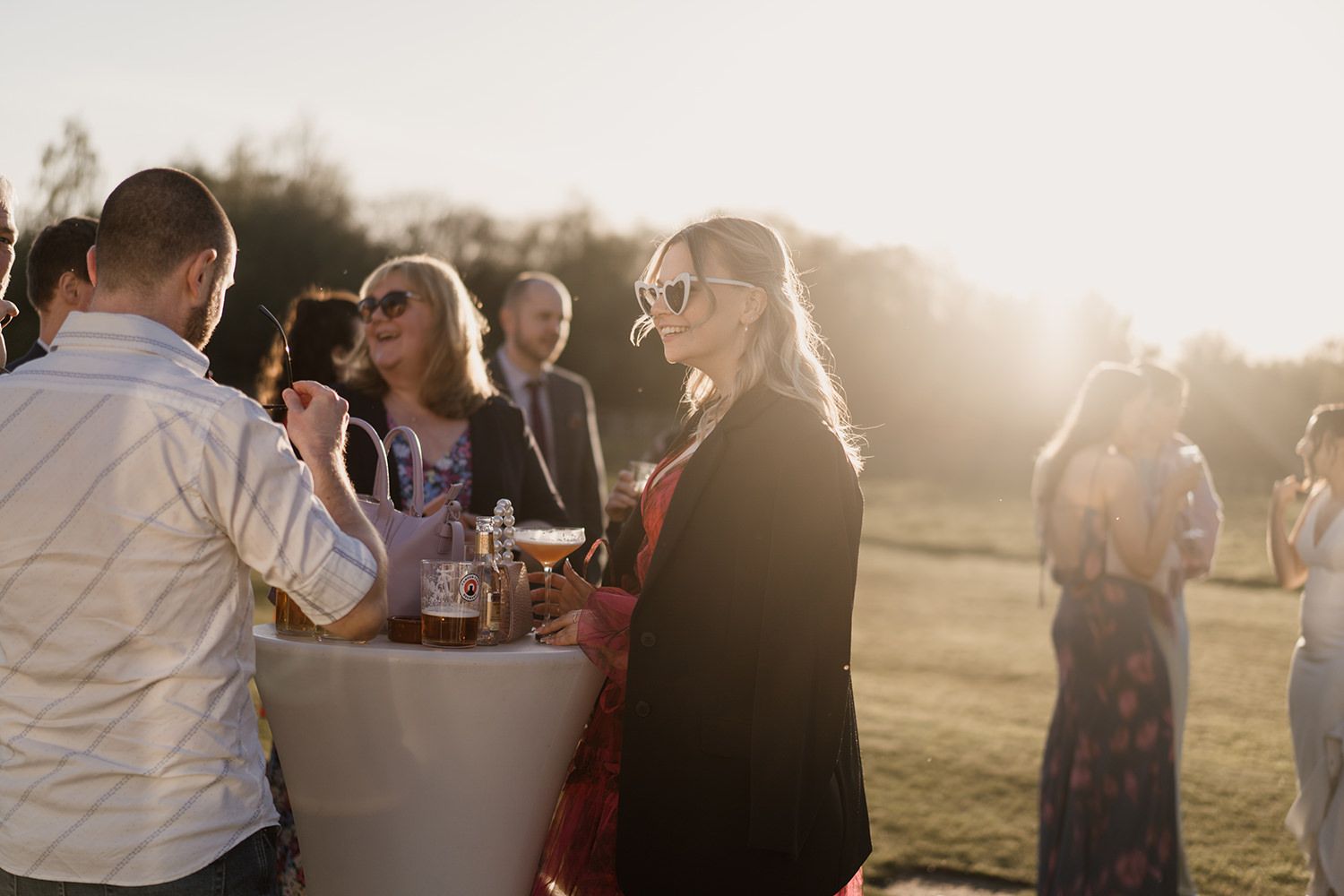 Wedding guests stood talking while standing on grass with the sun behind them.