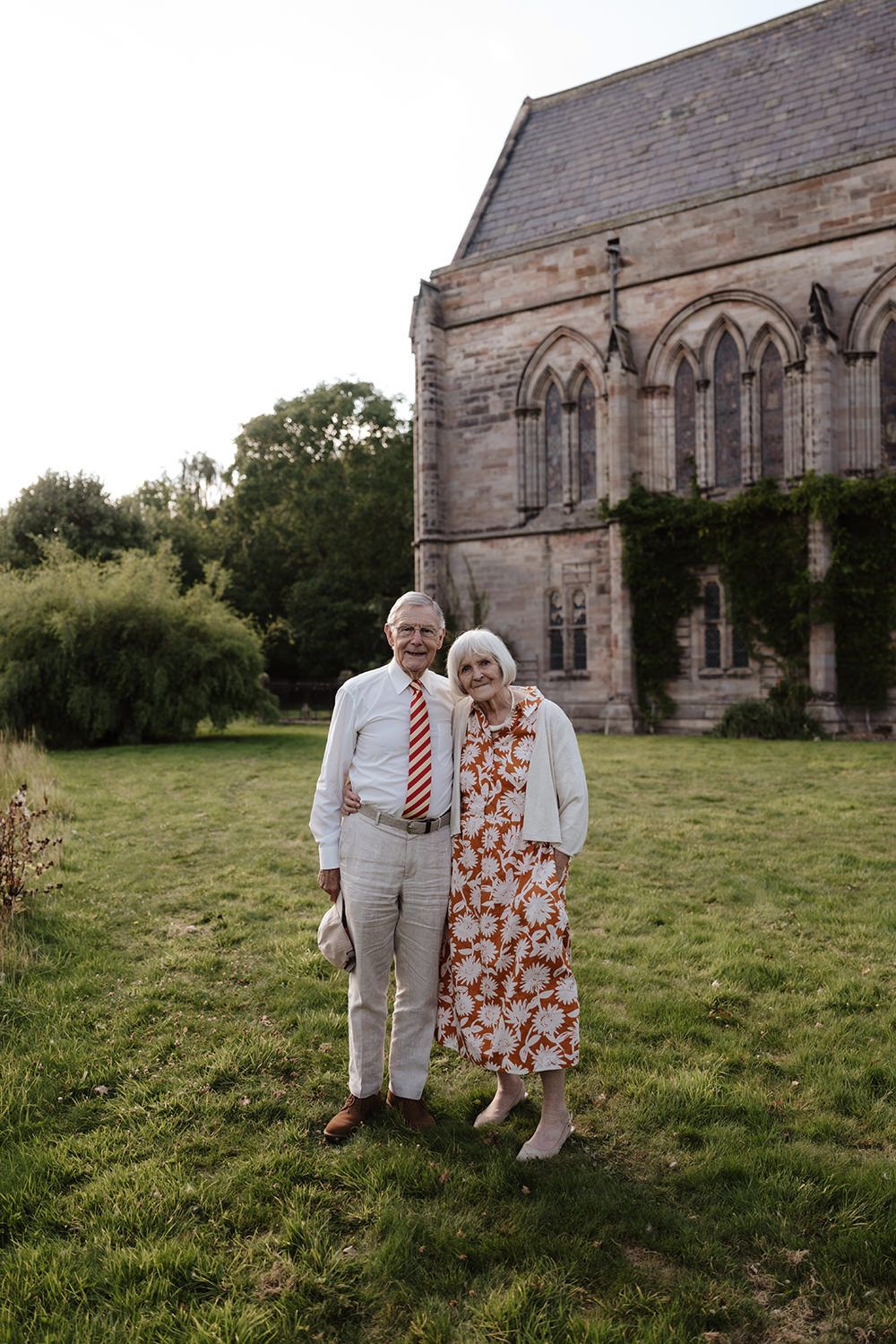 A couple shares an intimate moment together in a grassy field beside a stone church building.
