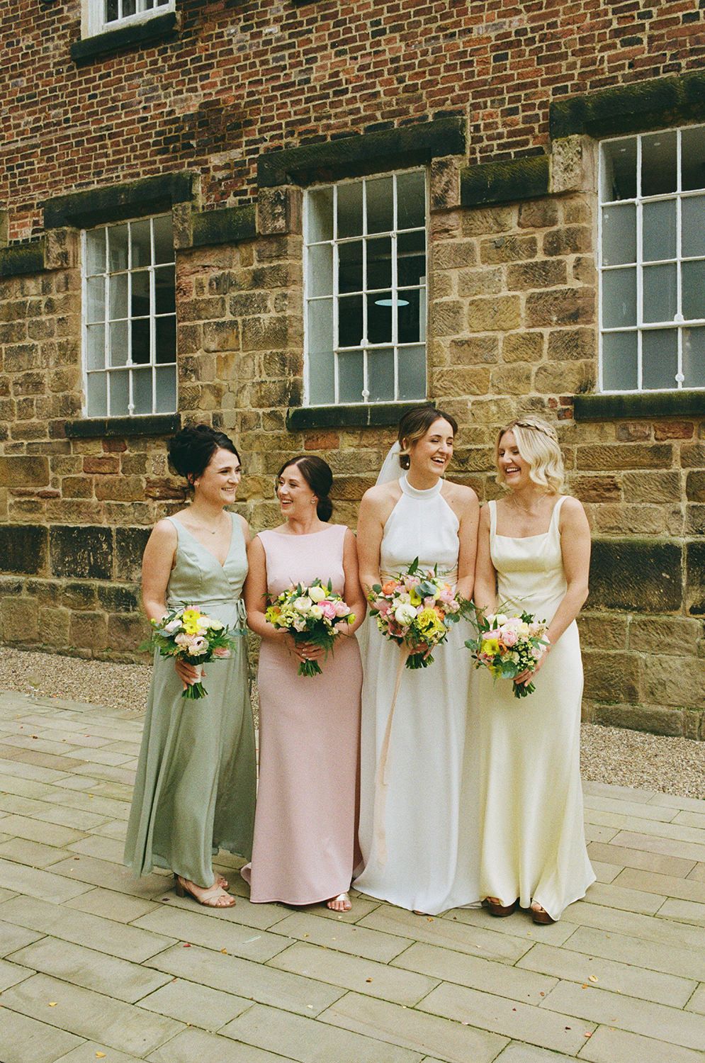 Bride and bridesmaids wearing pastel dresses smiling and laughing at The West Mill.