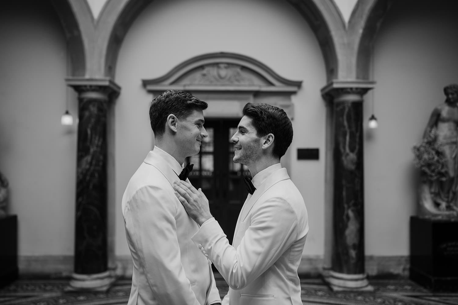 Two grooms holding hands smiling towards each other, they're wearing white blazers with a black bow tie.