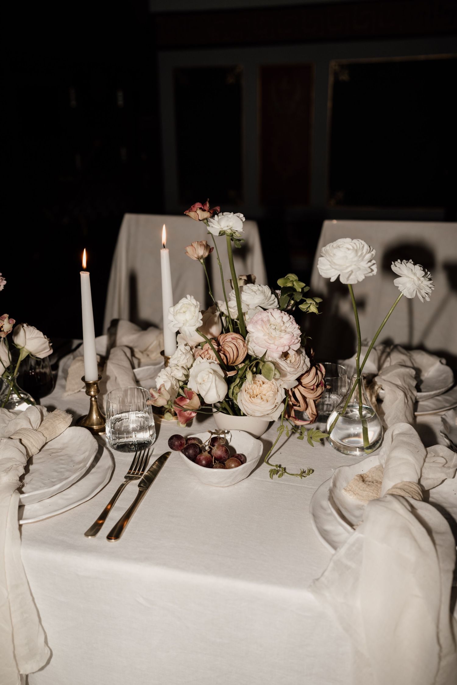 Elegant wedding table setting with white florals, candles, and gold accents on a white linen tablecloth.