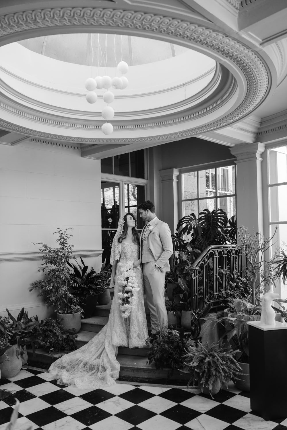 Black and white wedding photo of bride and groom standing under ornate ceiling in elegant venue with checkered floors.
