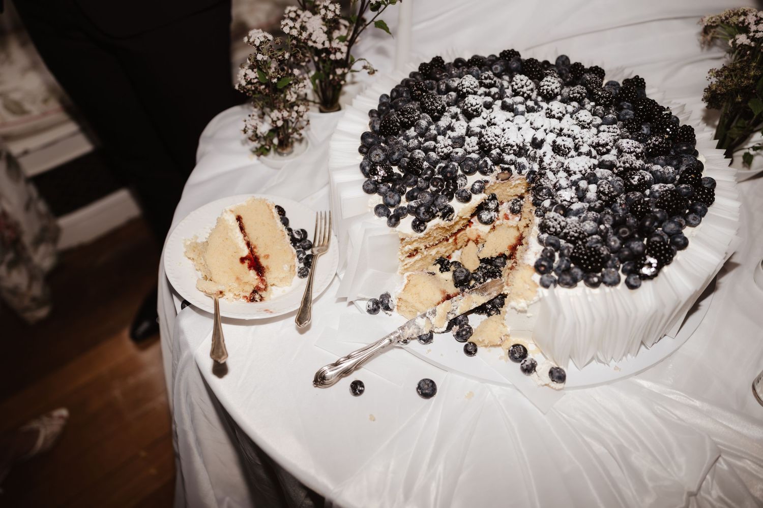 Wedding cake topped with fresh blueberries and powdered sugar with a slice served on a plate.