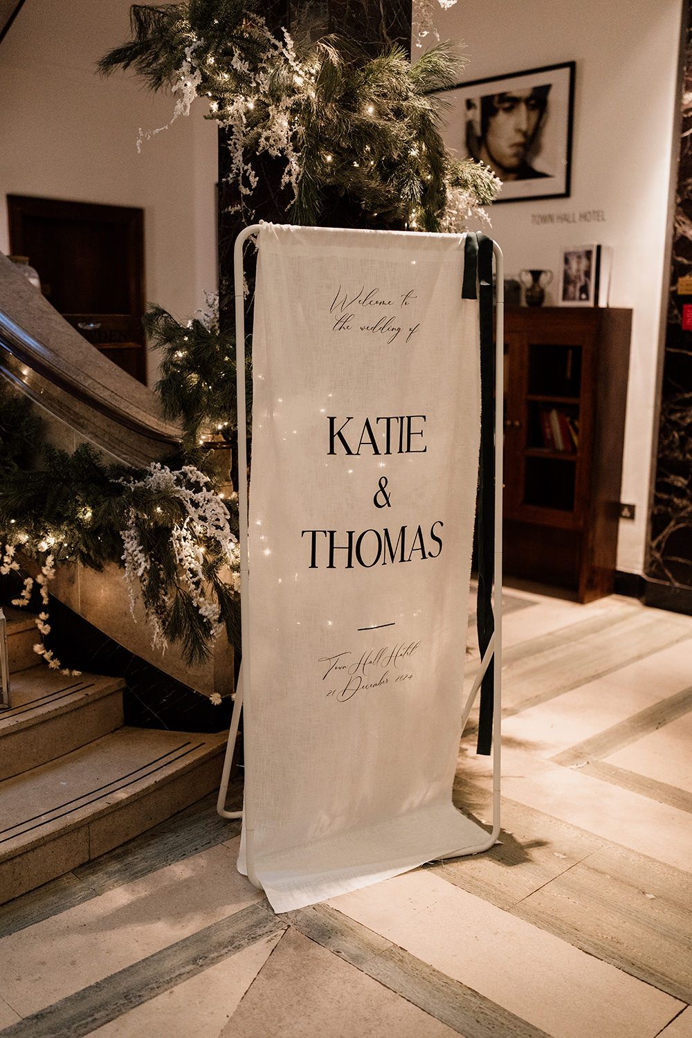 A white wedding welcome sign for Katie and Thomas stands beside stairs decorated with greenery and fairy lights.