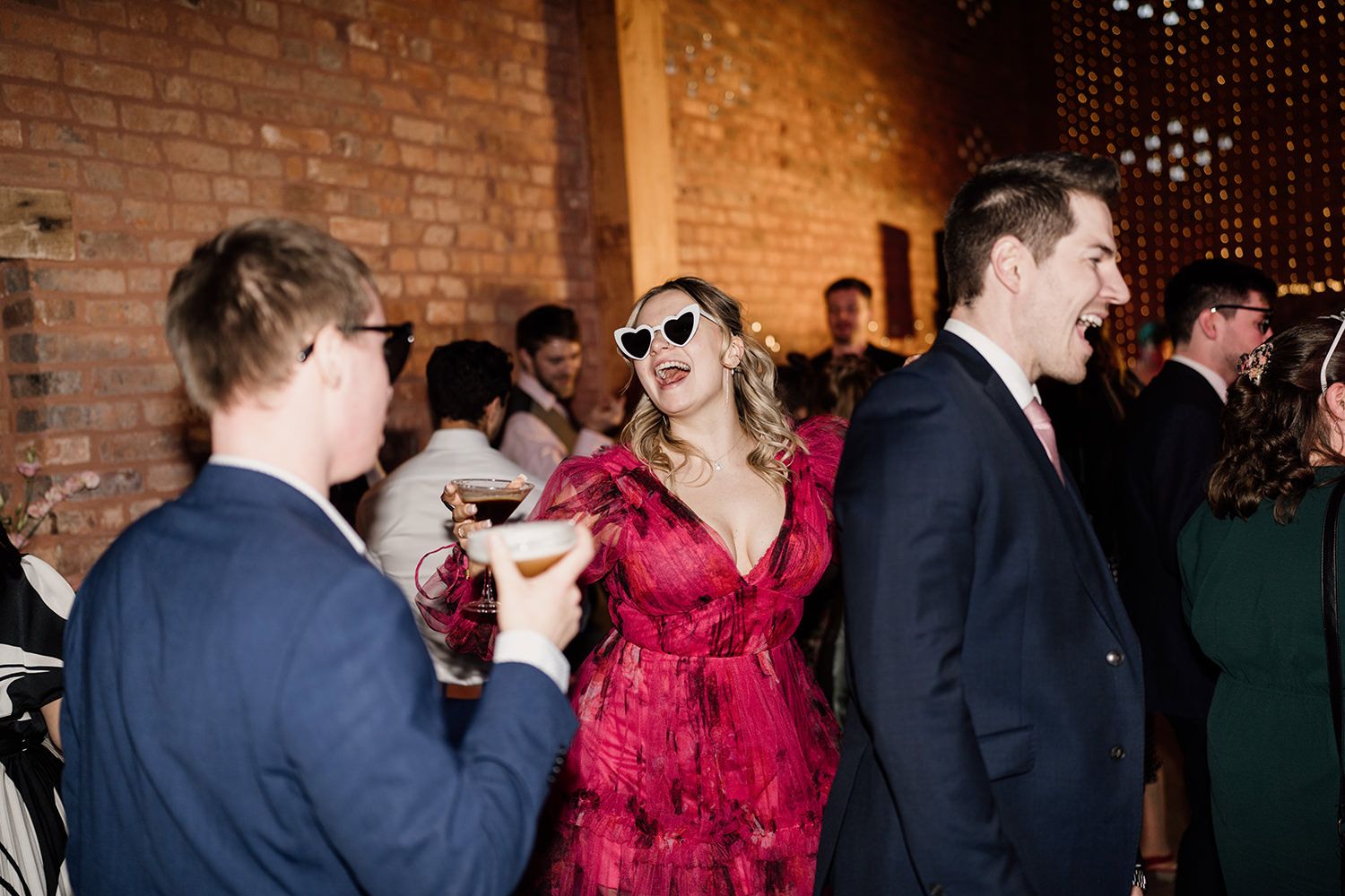 Wedding guest wearing a red dress and white sun glasses holding a cocktail while singing.