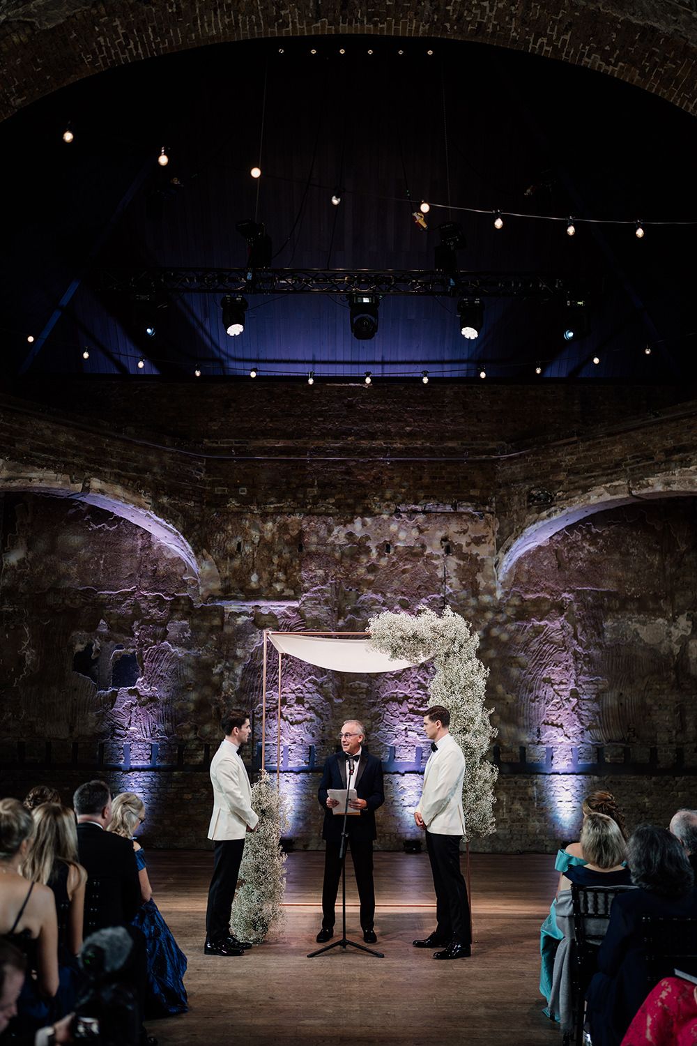 Two grooms stood during their wedding ceremony with cuppah and up lighters behind.