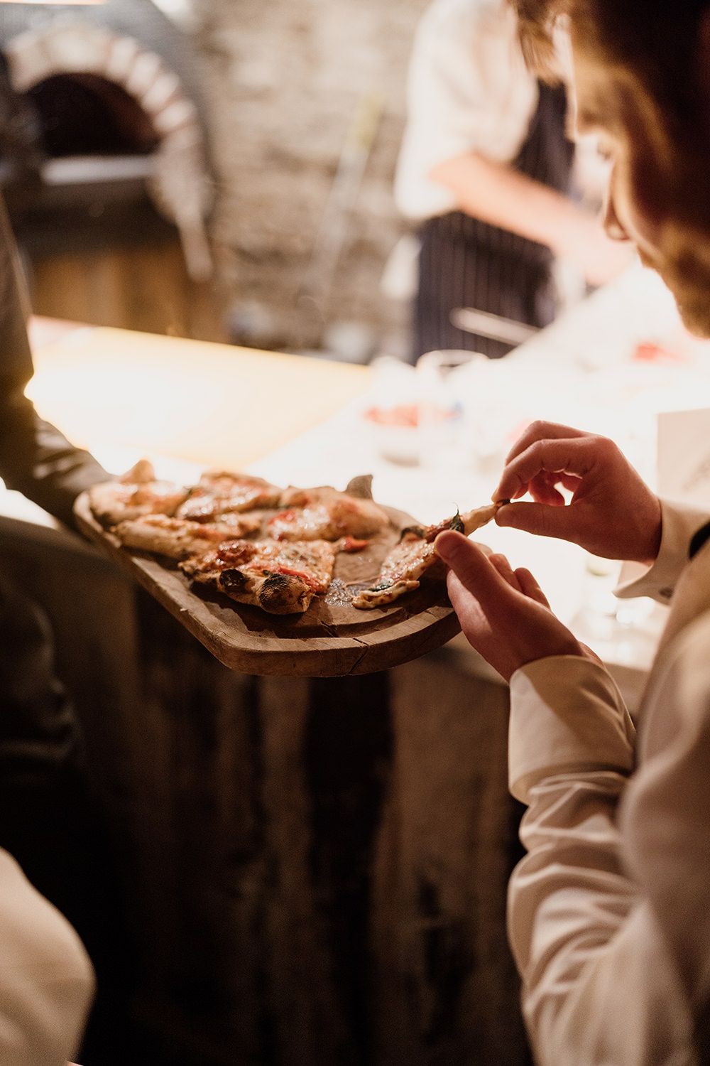 Person holding a wooden platter of hand made pizzas while someone takes a slice.