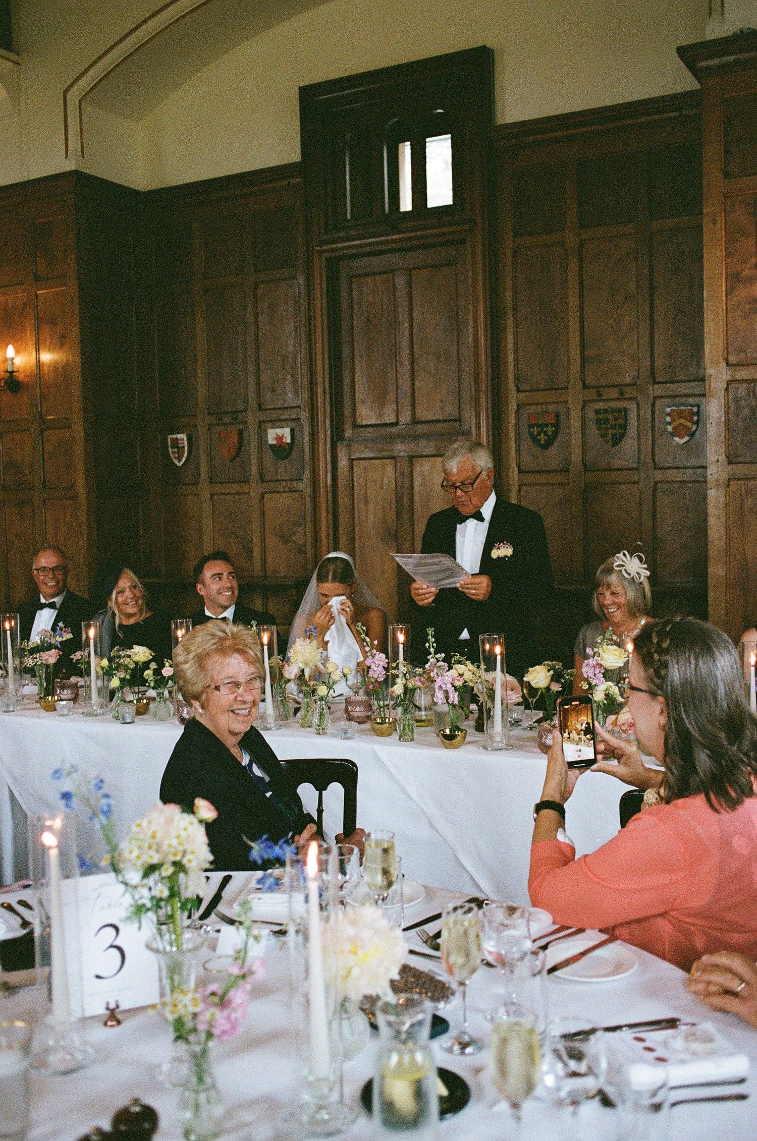 Wedding reception speech by man in tuxedo at head table with bride, guests, and floral centerpieces in elegant wood-paneled hall.