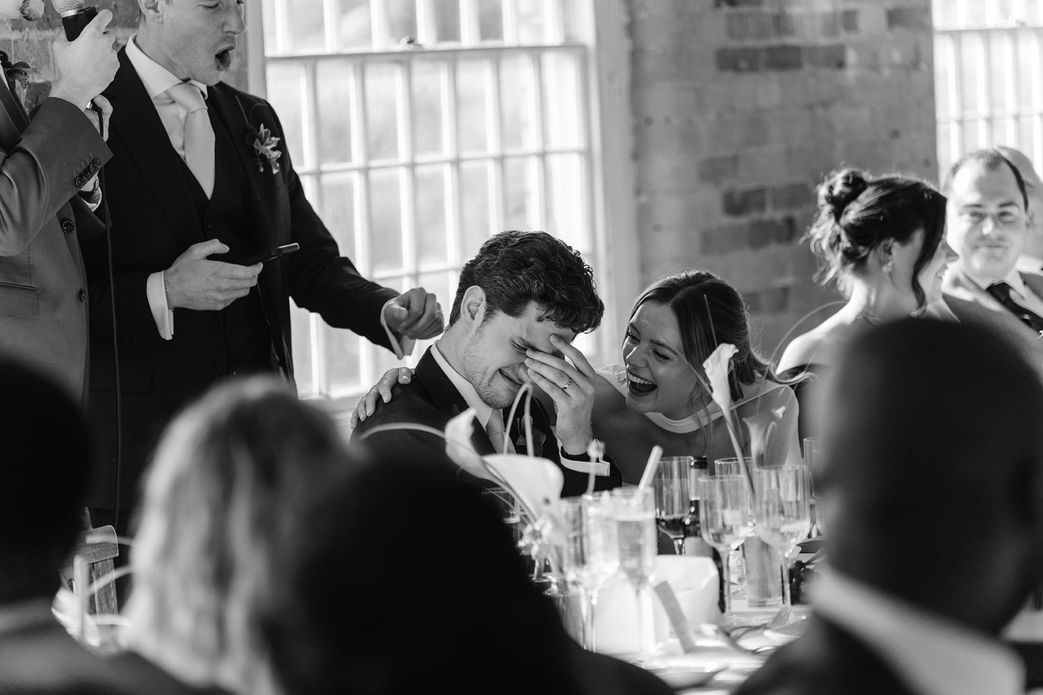 Black and white photo of bride and groom laughing together during wedding reception speeches.