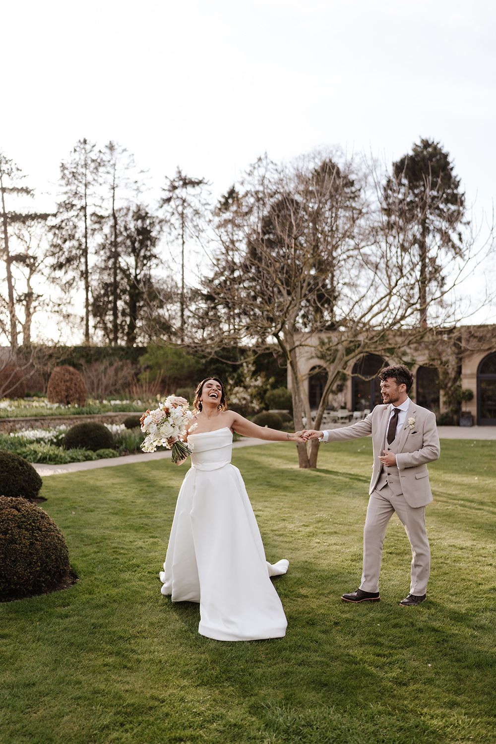 A couple celebrates joyfully on their wedding day in a garden setting with tall trees and manicured lawn.