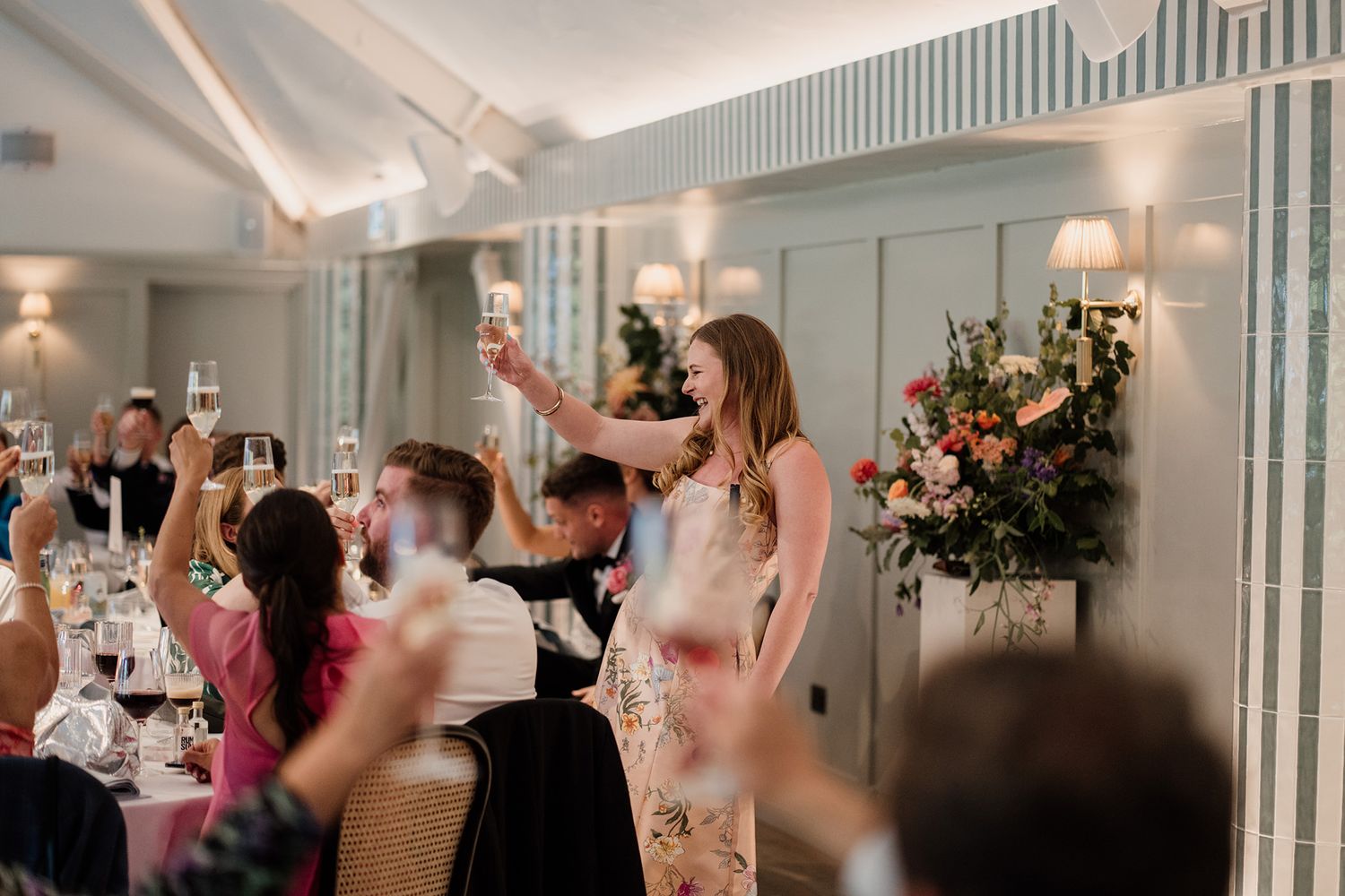 Guests raise their glasses in celebration during an elegant wedding reception at a brightly lit indoor venue.