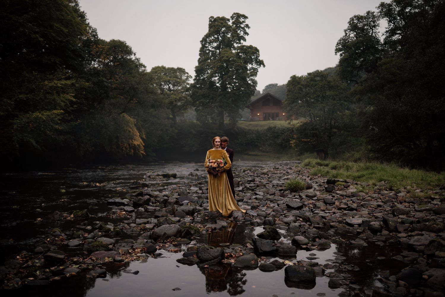 Couple stood amongst a dried river surrounded by trees wearing autumnal themed wedding clothes.