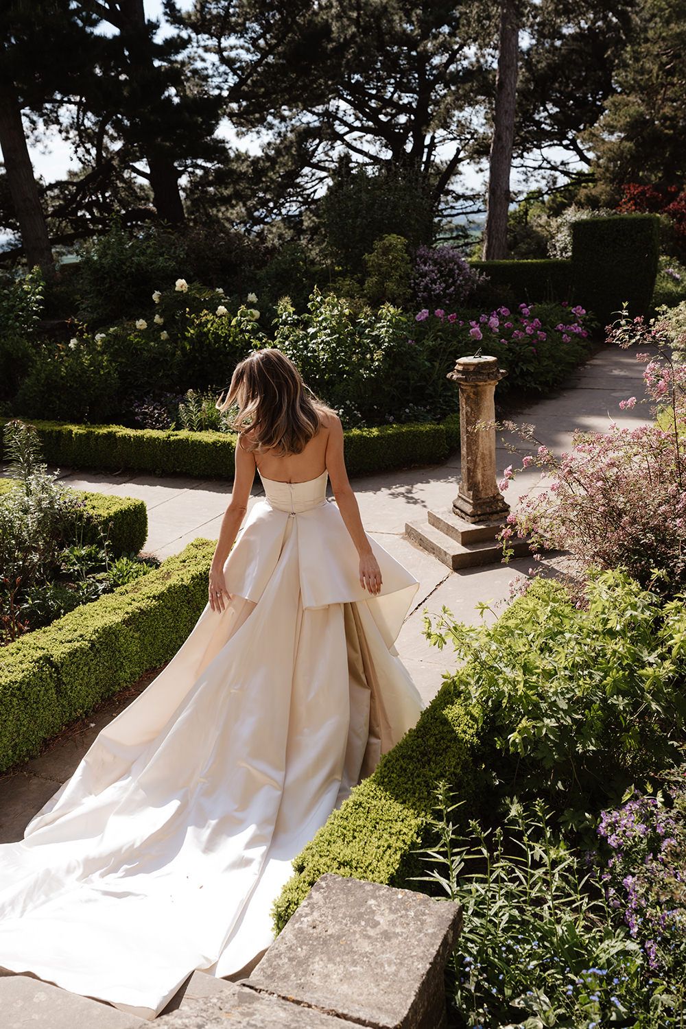 Beautiful wedding gown with flowing train photographed in a lush garden setting with stone pillars and blooming flowers.