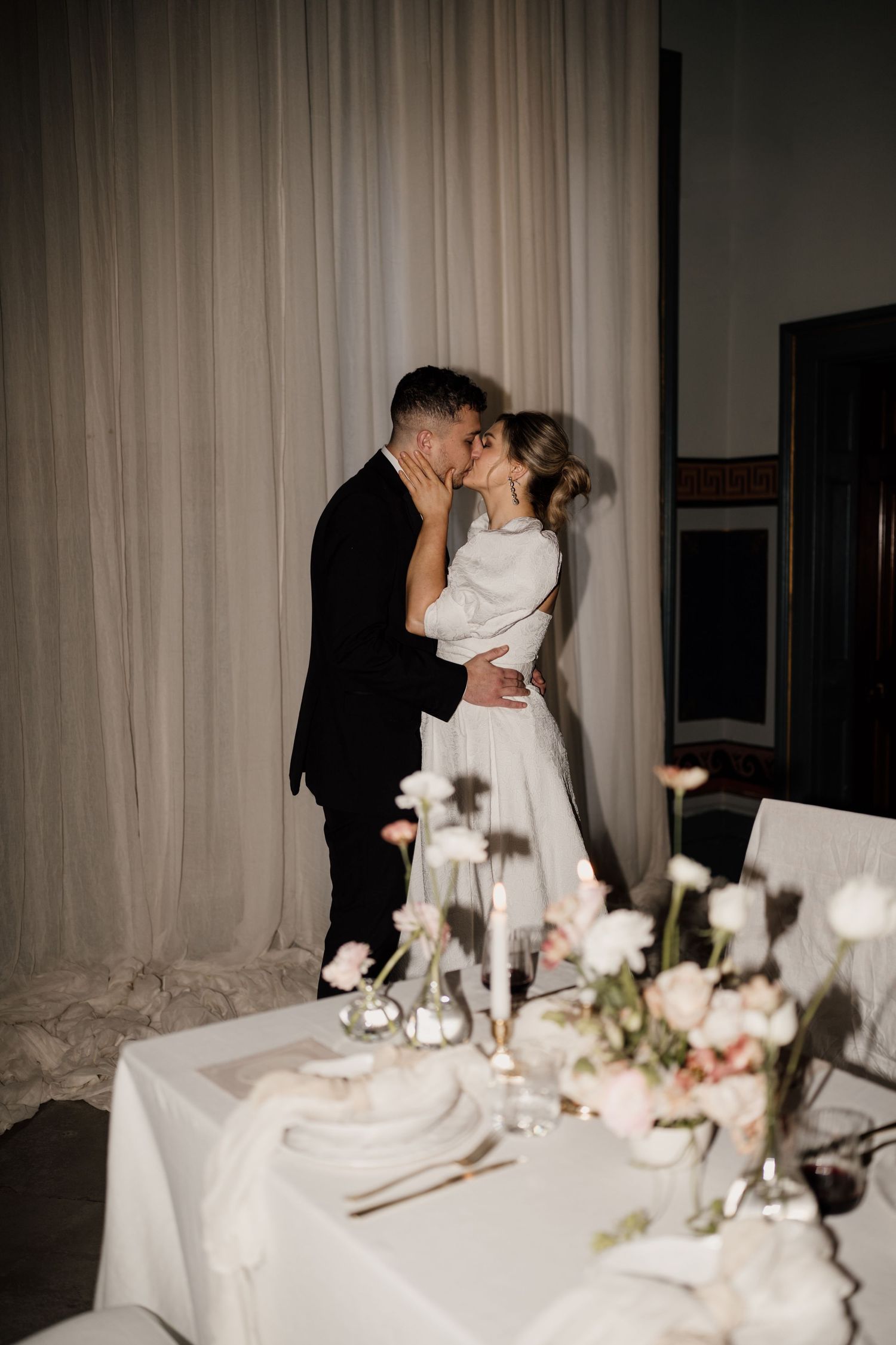 Bride and groom sharing a romantic kiss at elegant wedding reception with floral table setting and draped curtains.