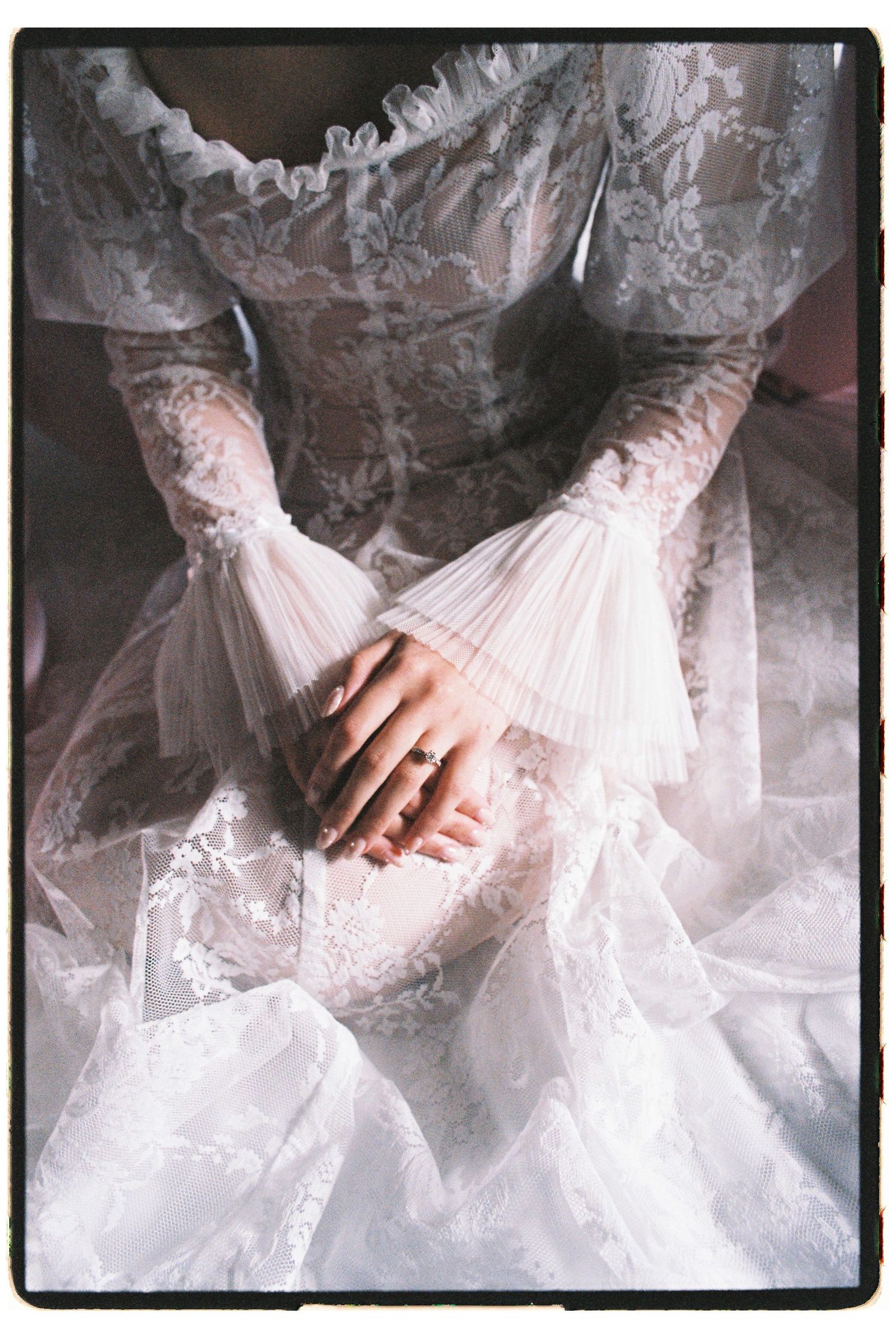 Close-up of a bride in a white lace wedding gown with long sleeves, hands folded gracefully in her lap.