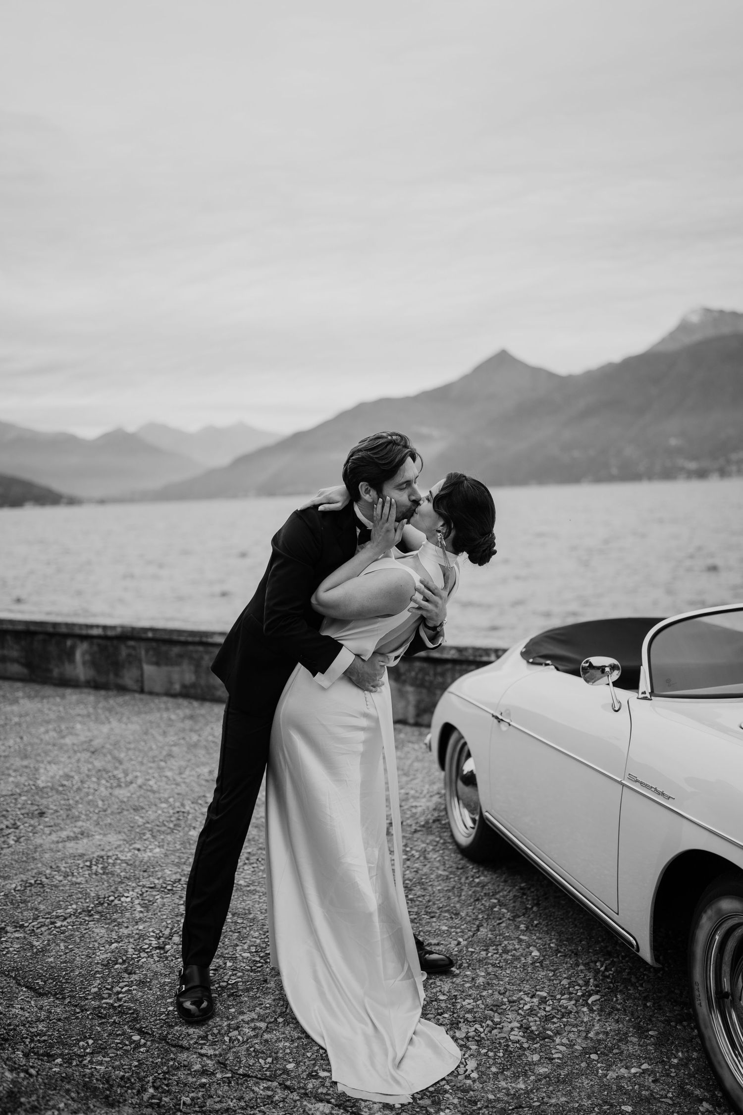 Black and white photo of groom dipping bride in elegant kiss beside vintage car with mountain lake view.