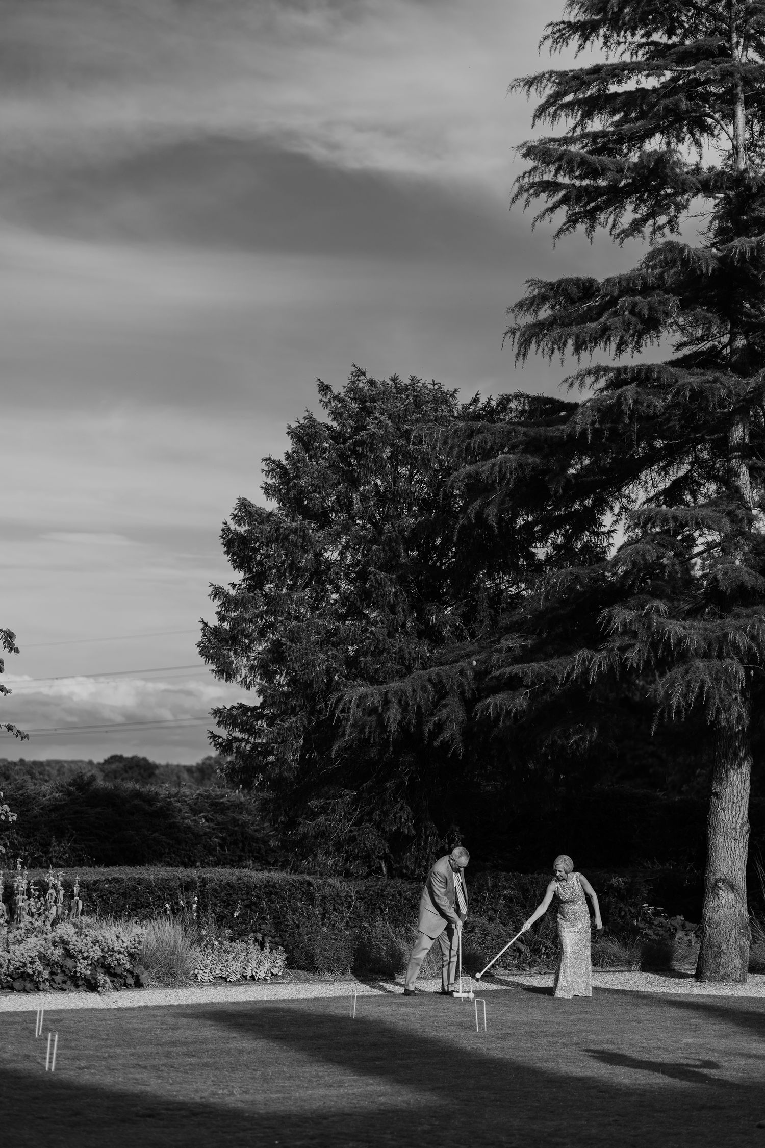 Black and white photo of two wedding guests playing croquet on lawn beside tall trees.