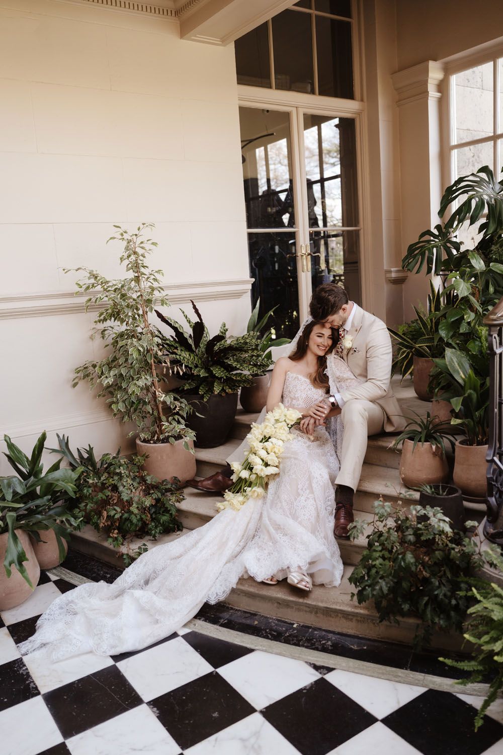 Bride and groom sitting on steps surrounded by lush potted plants at elegant venue with checkered tile floor.