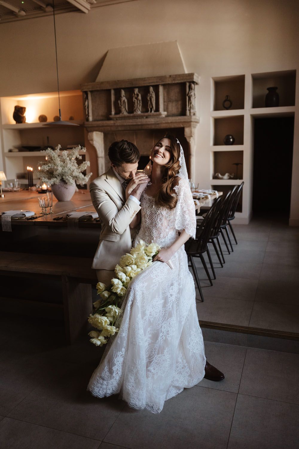 Bride in lace gown holds yellow tulips as groom kisses her hand in elegant rustic venue with stone fireplace.