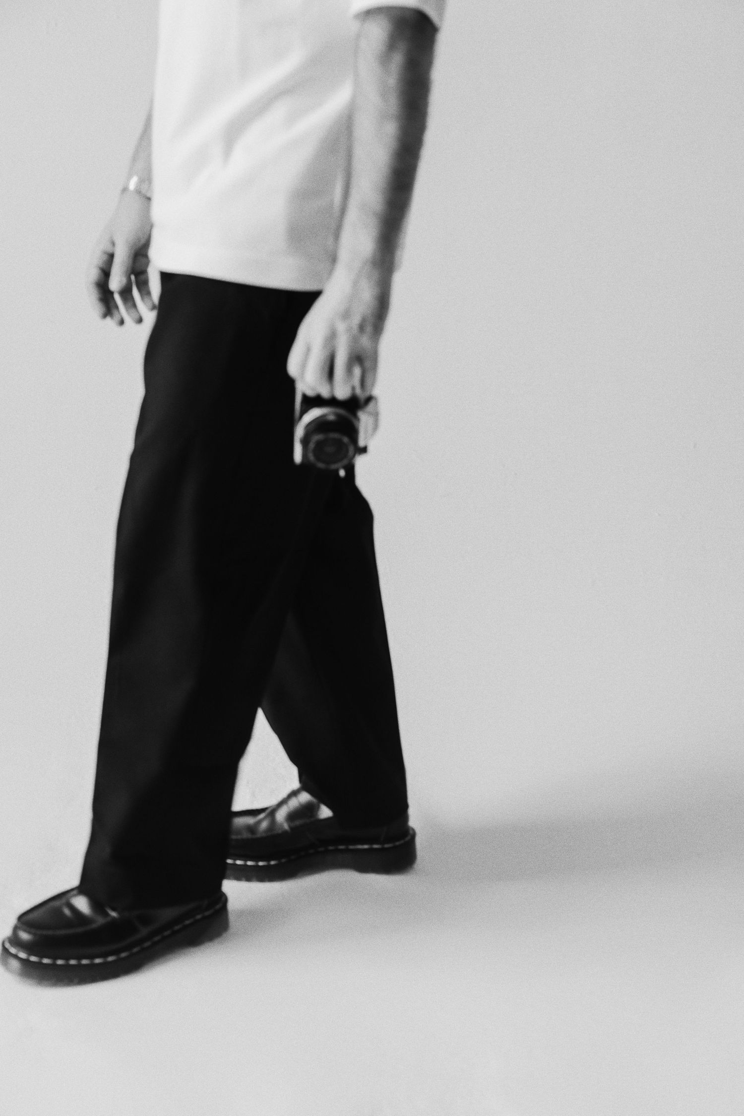 Black and white photo of a person in wide-leg black pants and Dr. Martens holding a film camera.