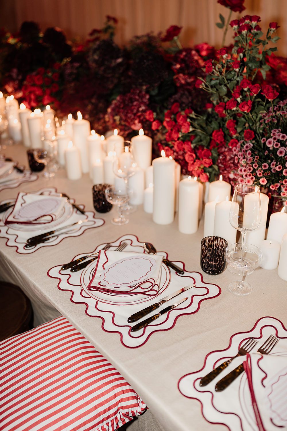 Elegant table setting with white candles and red flowers featuring striped cushions and decorative plate settings.