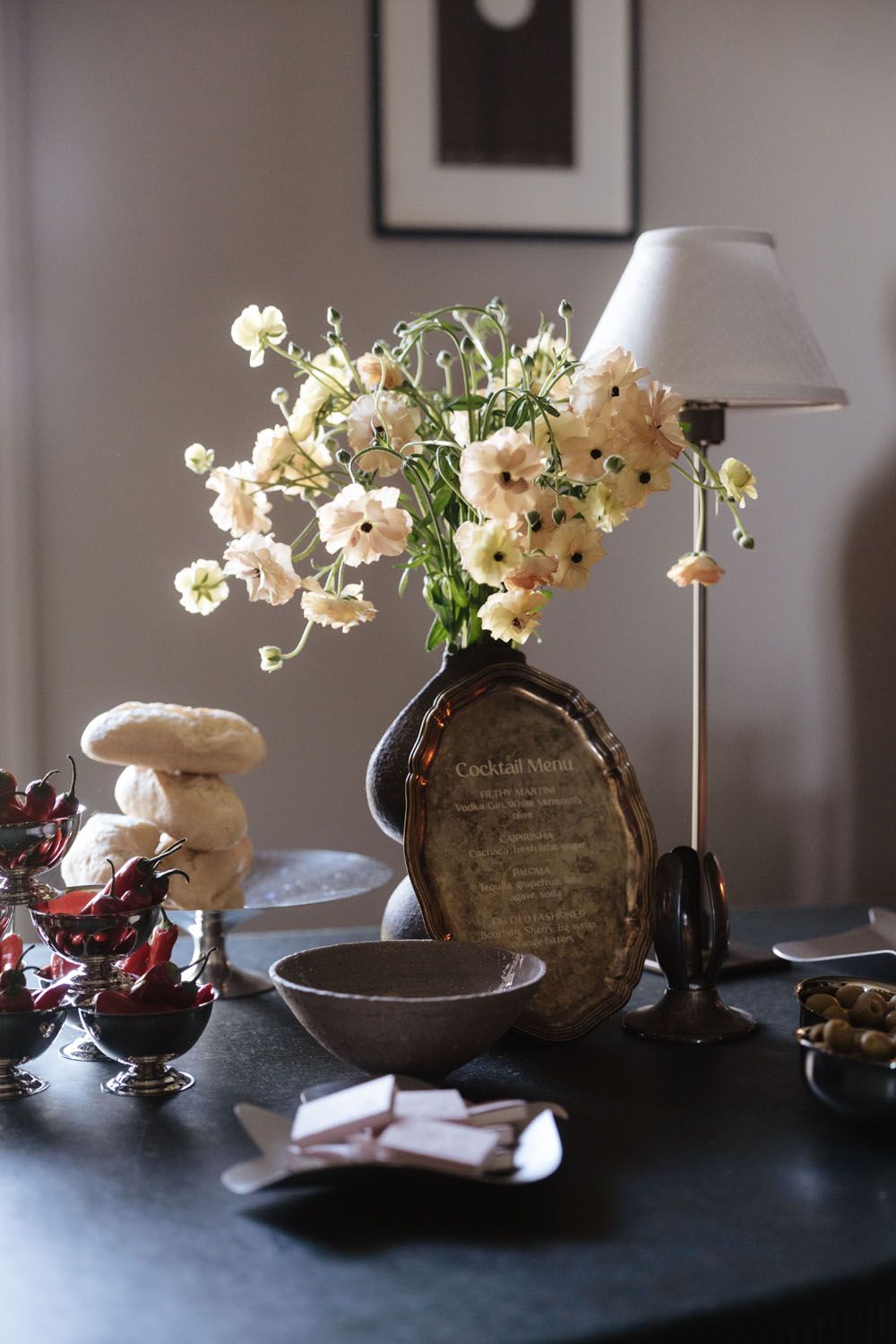 Elegant table display with white flowers in dark vase, cocktail menu, lamp, cherry desserts, and decorative bowl.