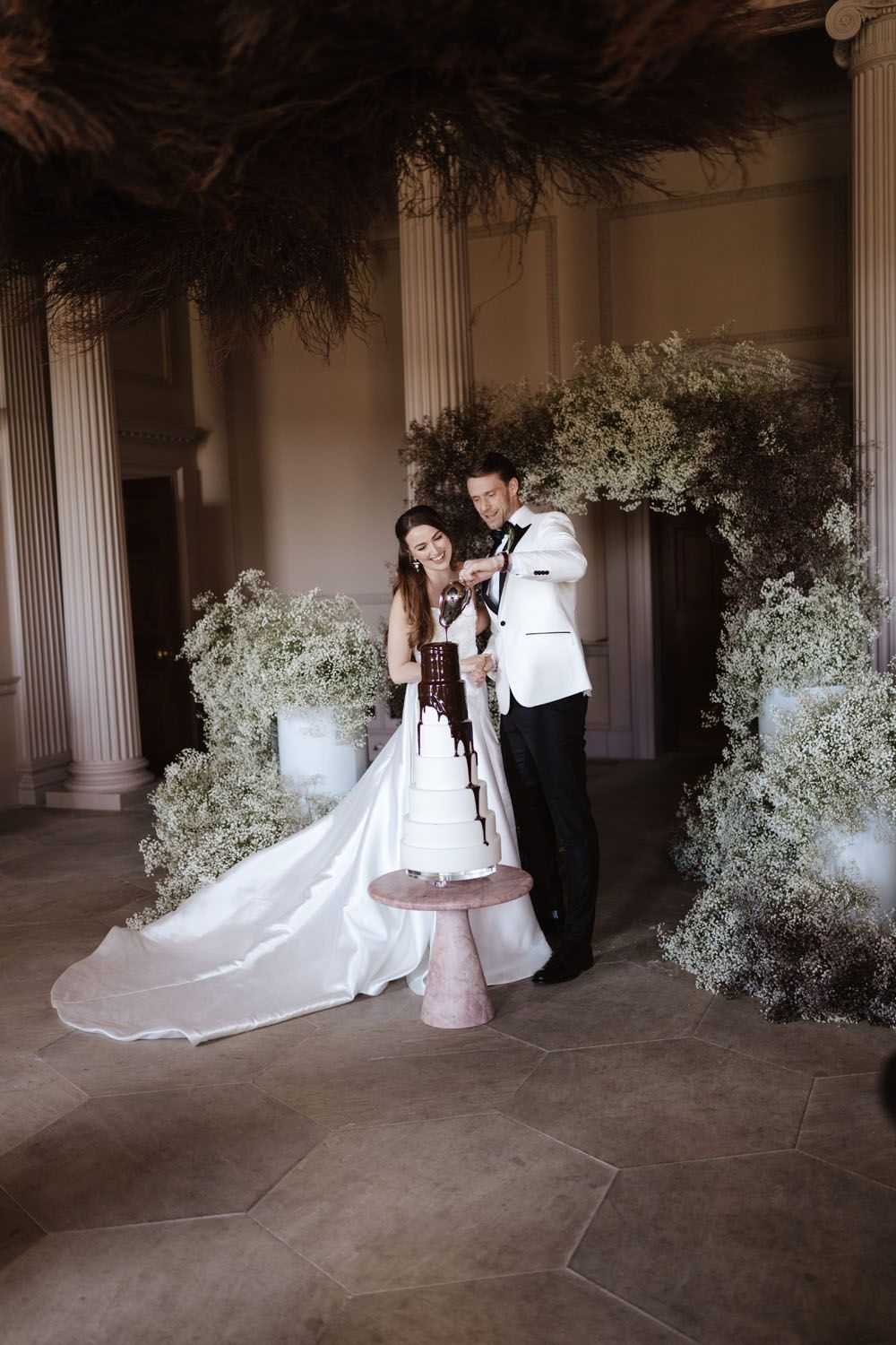 Bride and groom cutting a chocolate drip wedding cake surrounded by lush greenery and floral arrangements in an elegant venue.