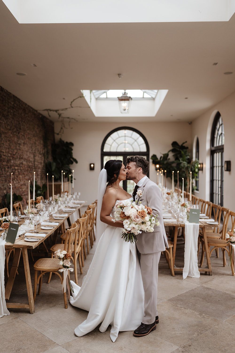 A couple embraces in an elegant wedding reception venue with long tables, skylight, and exposed brick wall.