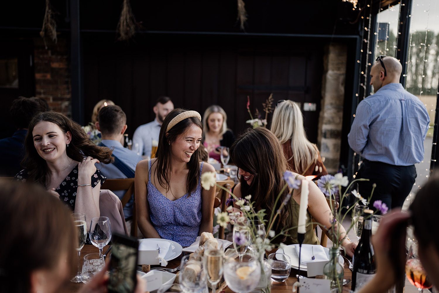 Wedding guest wearing a purple dress and white pearl headband smiling and laughing while sat during wedding speeches.