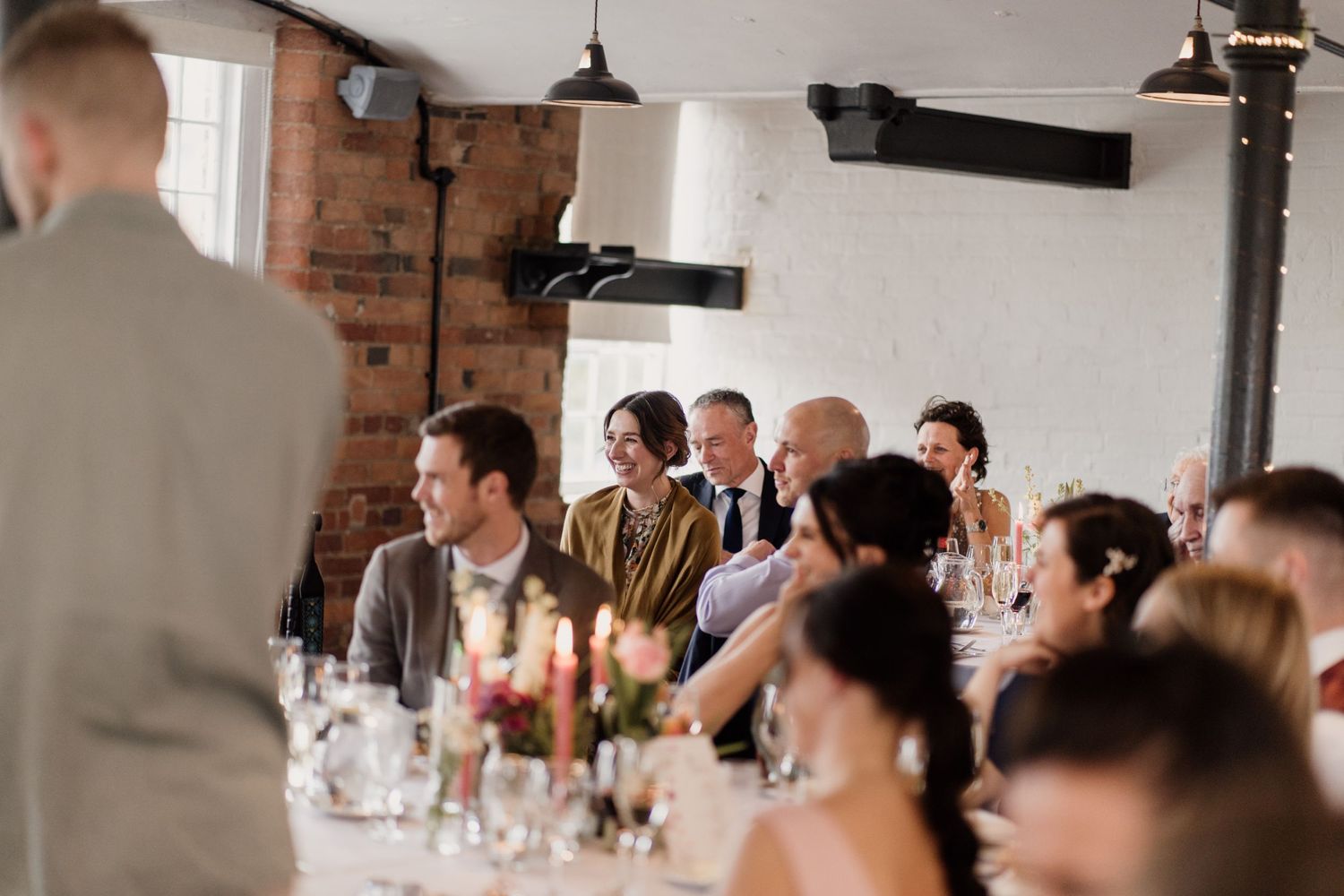 Wedding reception guests seated at long tables in rustic brick venue with elegant table settings and lighting.
