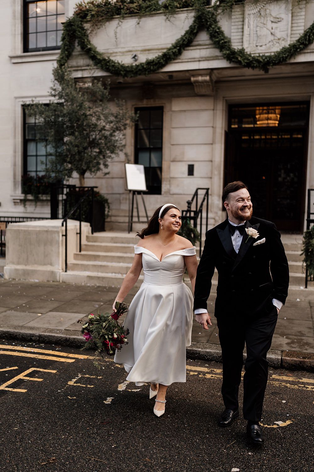 A couple walks together on a rainy street outside a historic building decorated with holiday garlands and greenery.