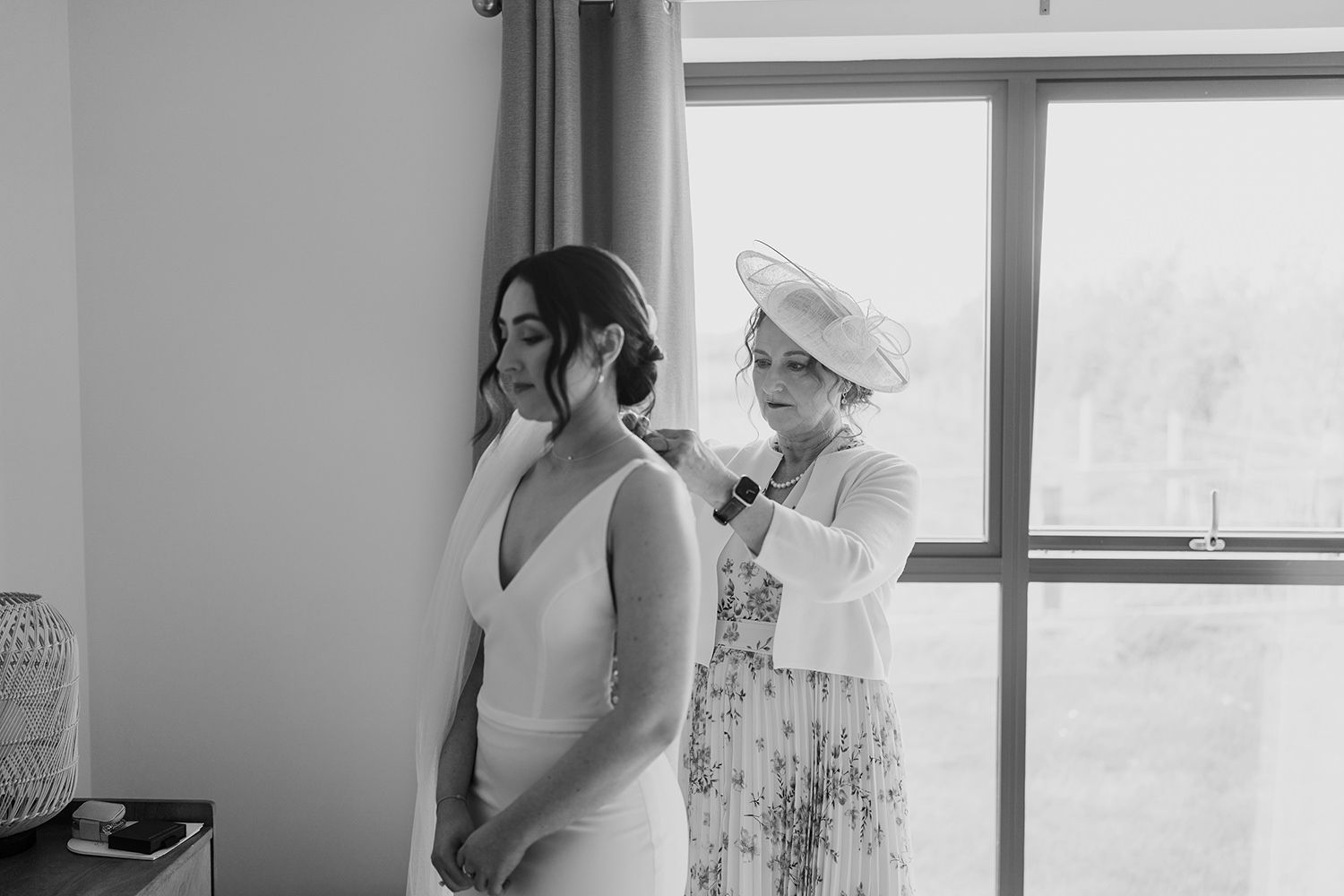Bride facing camera as mother stands behind her and fastens her necklace.