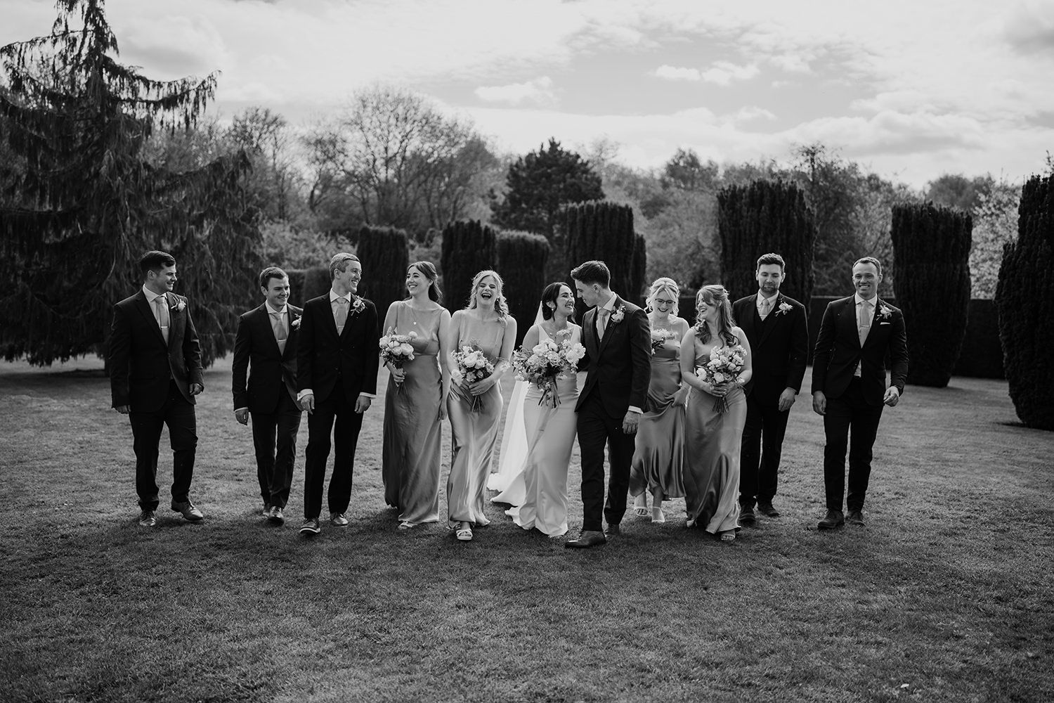 Groomsmen and Bridesmaids walking in a line laughing and talking through the gardens at The Holford Estate.