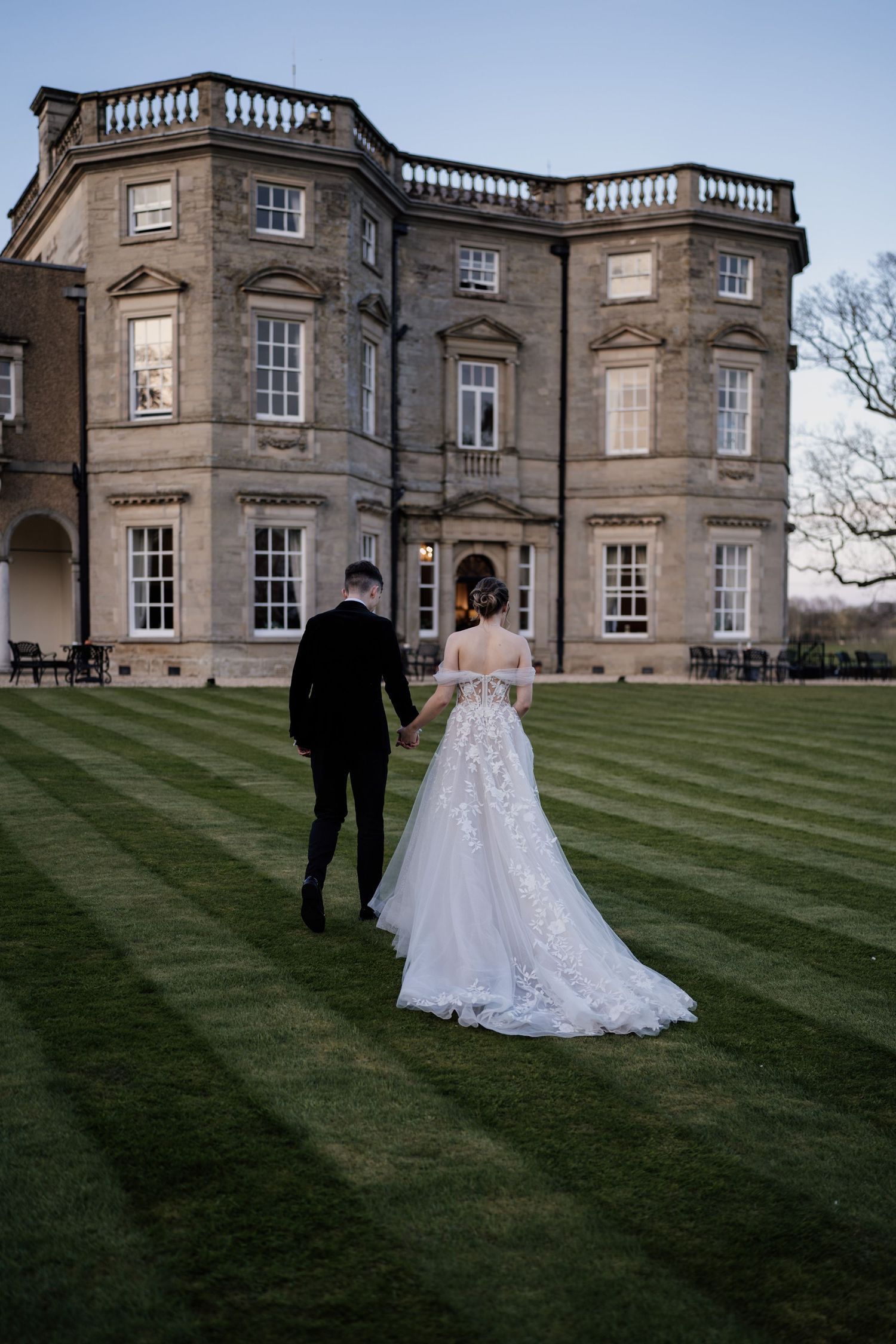 Bride and groom holding hands and walking towards Bourton Hall with blue skies on their wedding day.