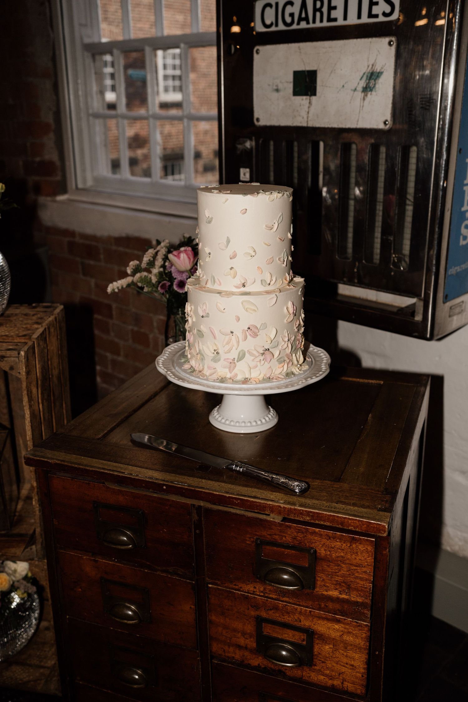 Two-tiered white wedding cake with delicate floral details displayed on vintage wooden drawer unit.