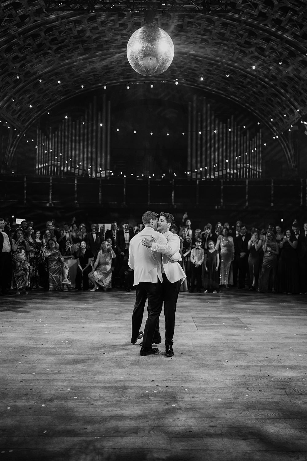 Two grooms performing their first dance with guests stood in a line watching and a disco ball above.