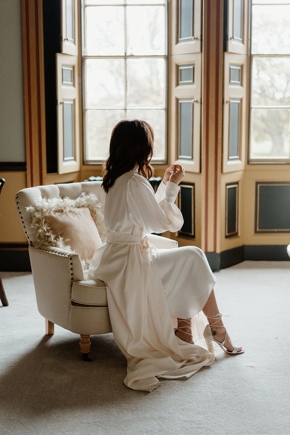 Bride sat in chair looking out of a grand victorian window.