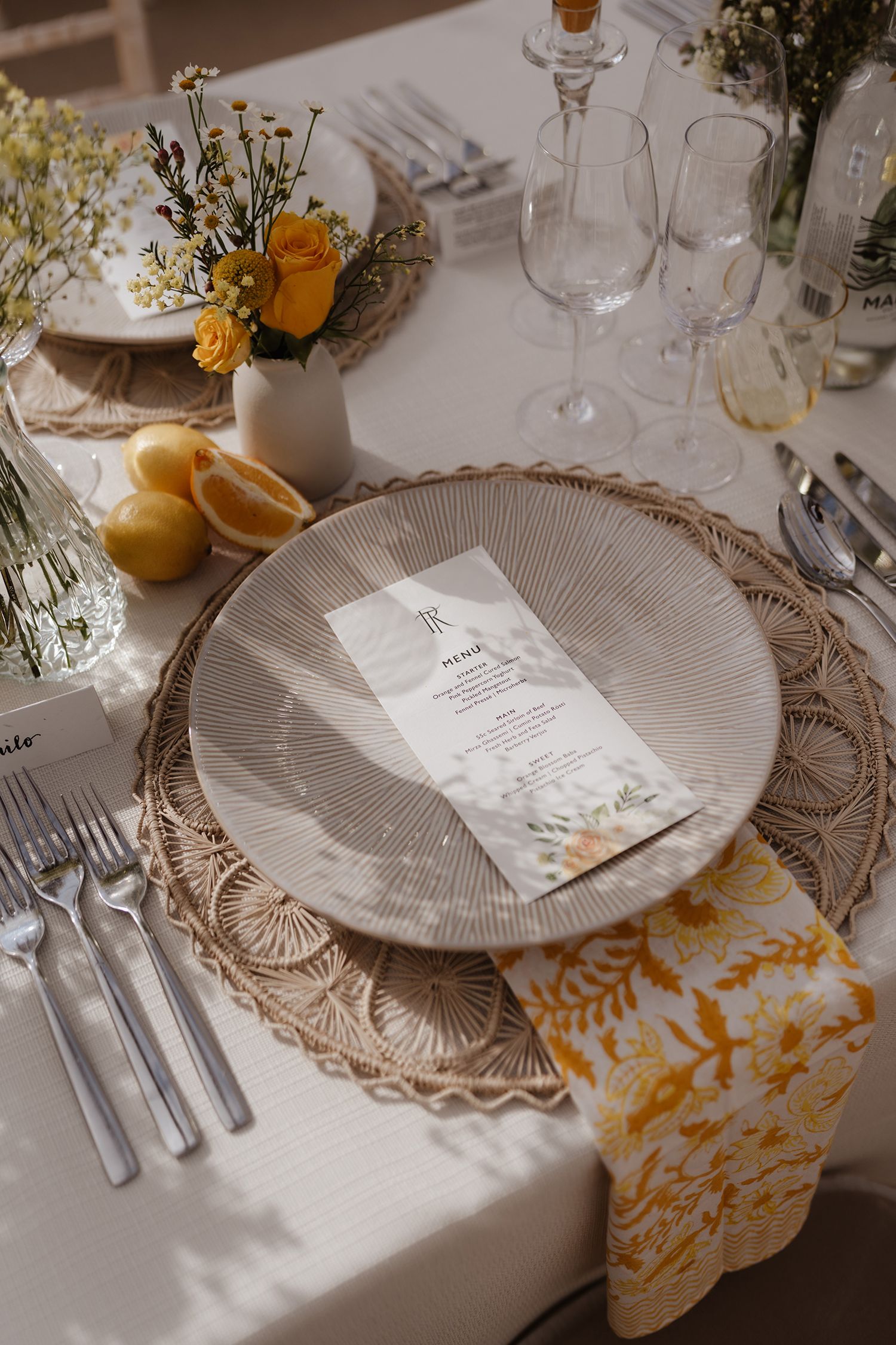 Elegant wedding place setting with woven charger, menu card, yellow roses and fresh lemons.