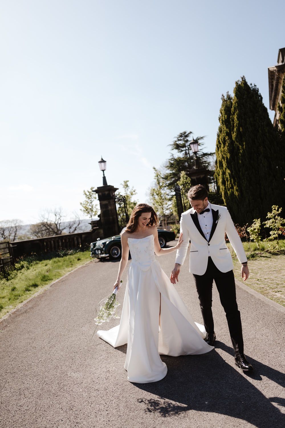 Bride in white gown and groom in white tuxedo walking hand in hand on a sunlit path at an elegant wedding venue.